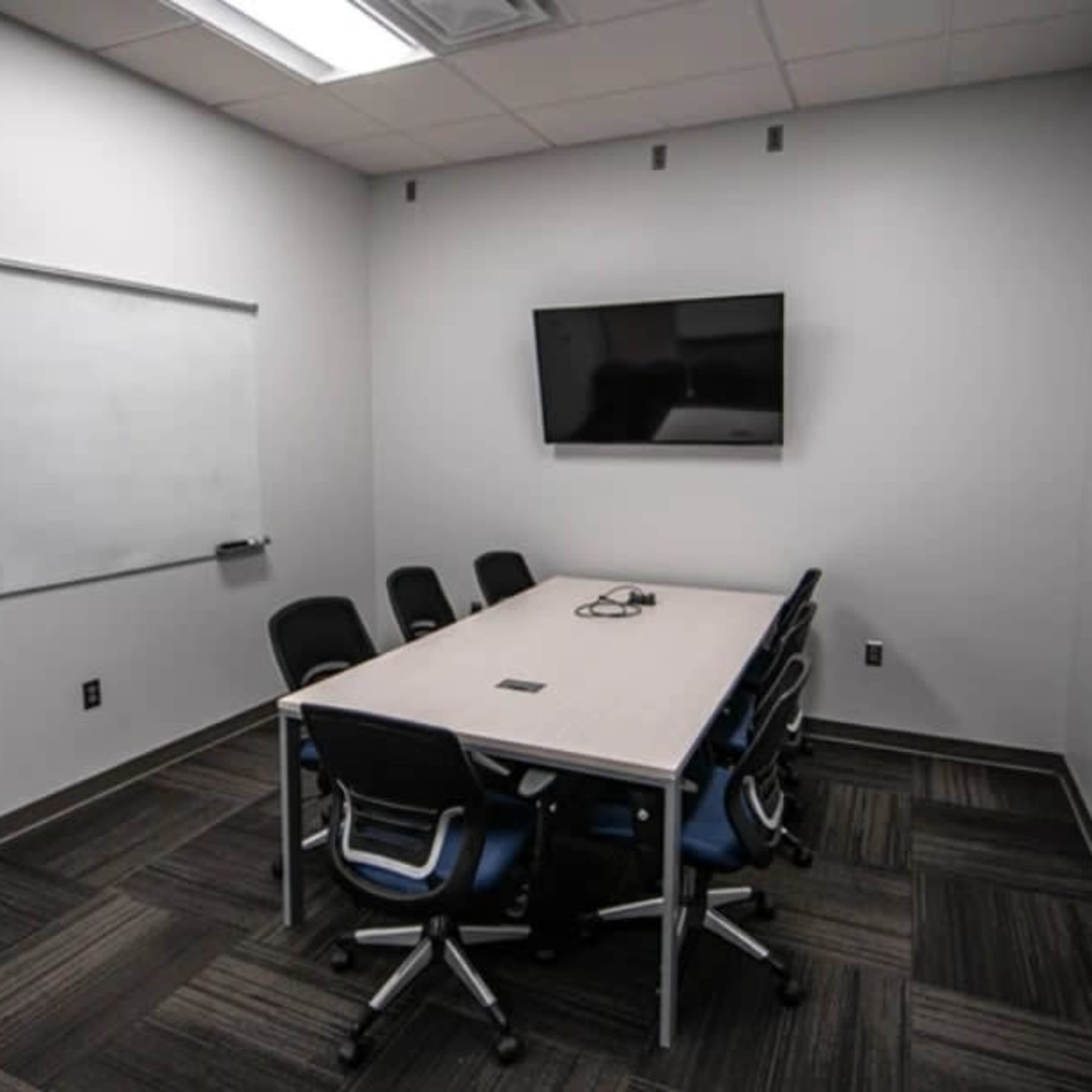 A small conference room features a whiteboard, a wall-mounted television, and a rectangular table surrounded by six chairs.