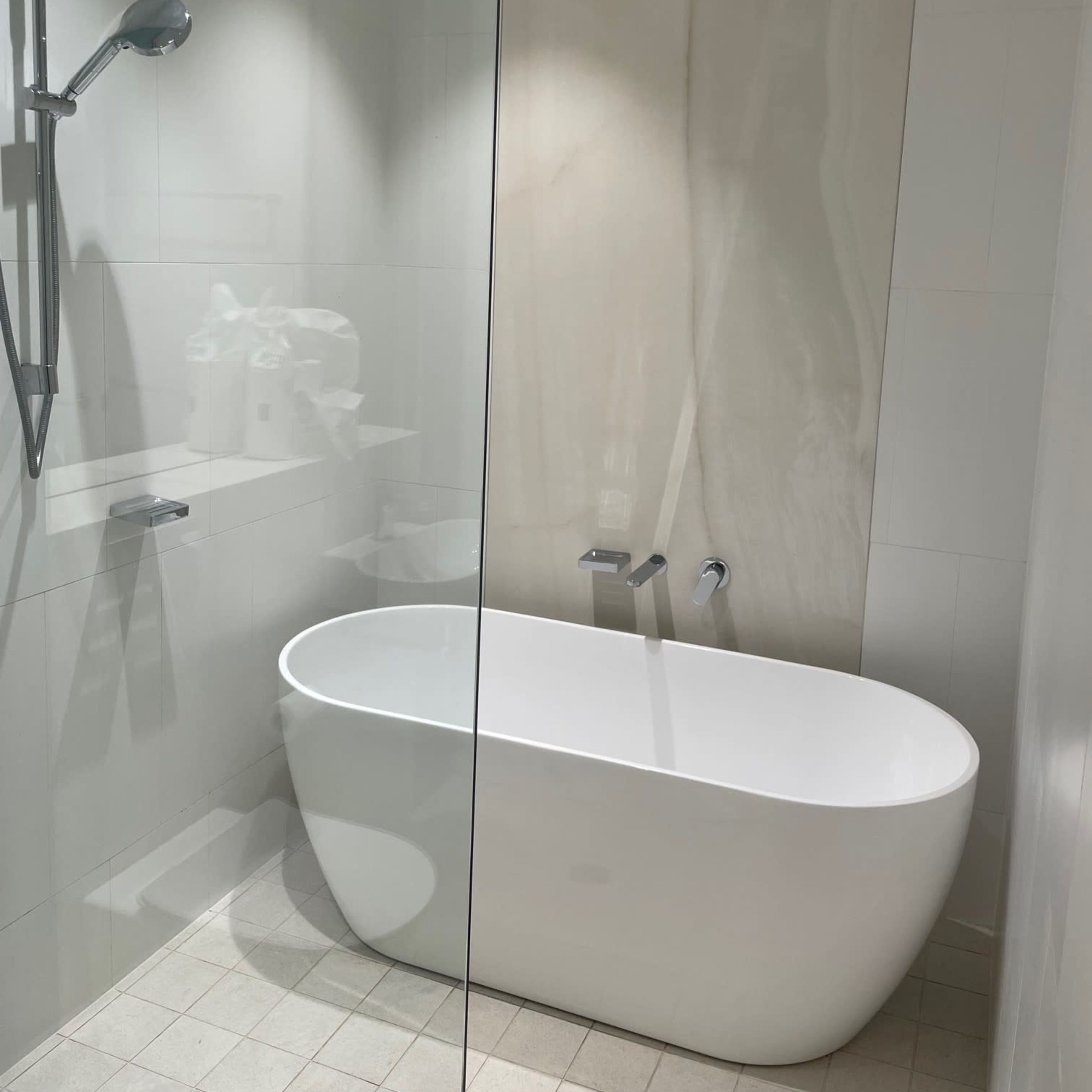 A modern bathroom featuring a freestanding white bathtub, a shower area with a glass partition, and light-colored tiled walls.