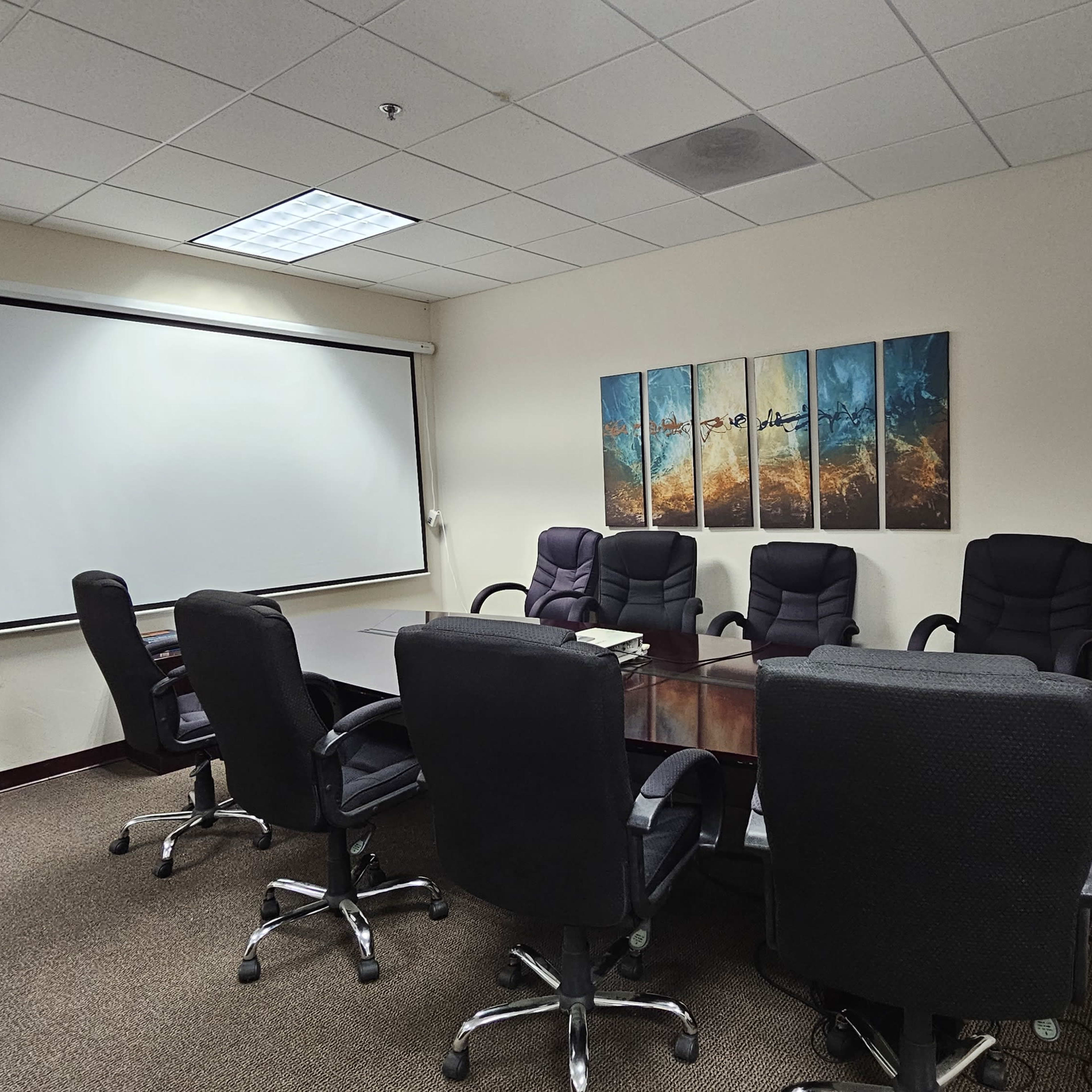 A conference room features a large table surrounded by several black office chairs, with a projector screen and artworks on the walls.