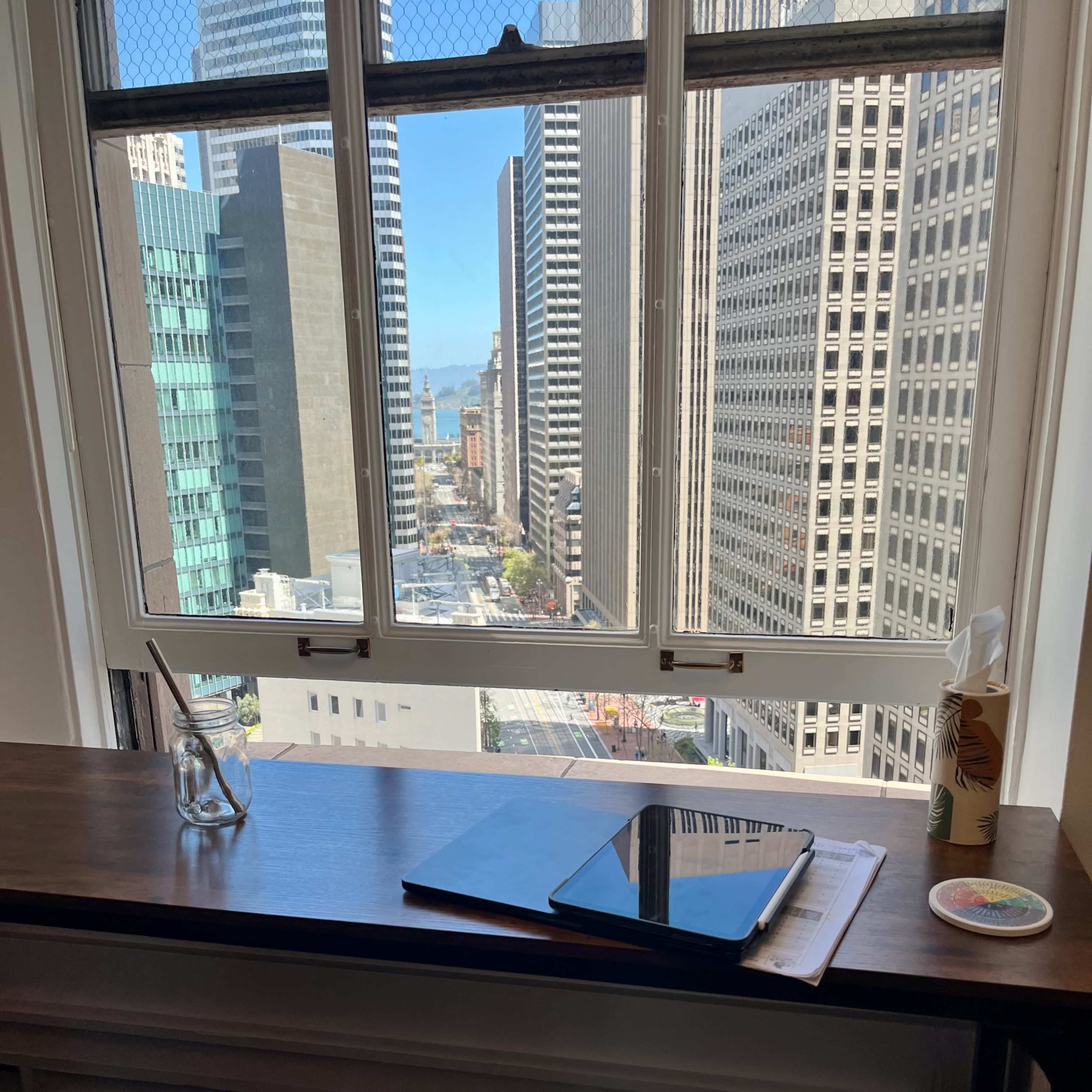 A workspace with a notebook and pen sits adjacent to a large window overlooking a cityscape with tall buildings and a street below.