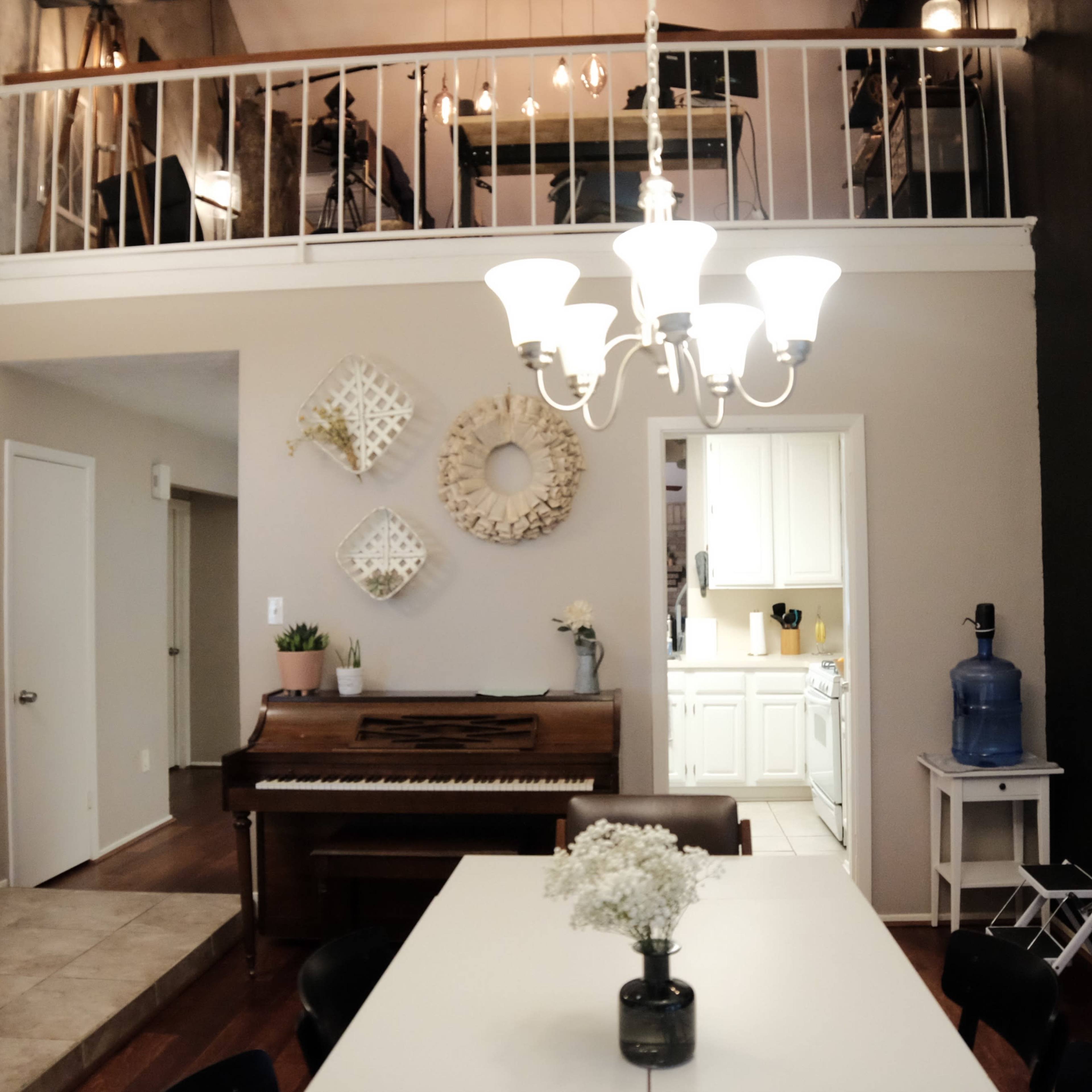 A modern dining area with a white table, black chairs, a piano, and decor on the walls, leading to an open kitchen and a lofted space above.