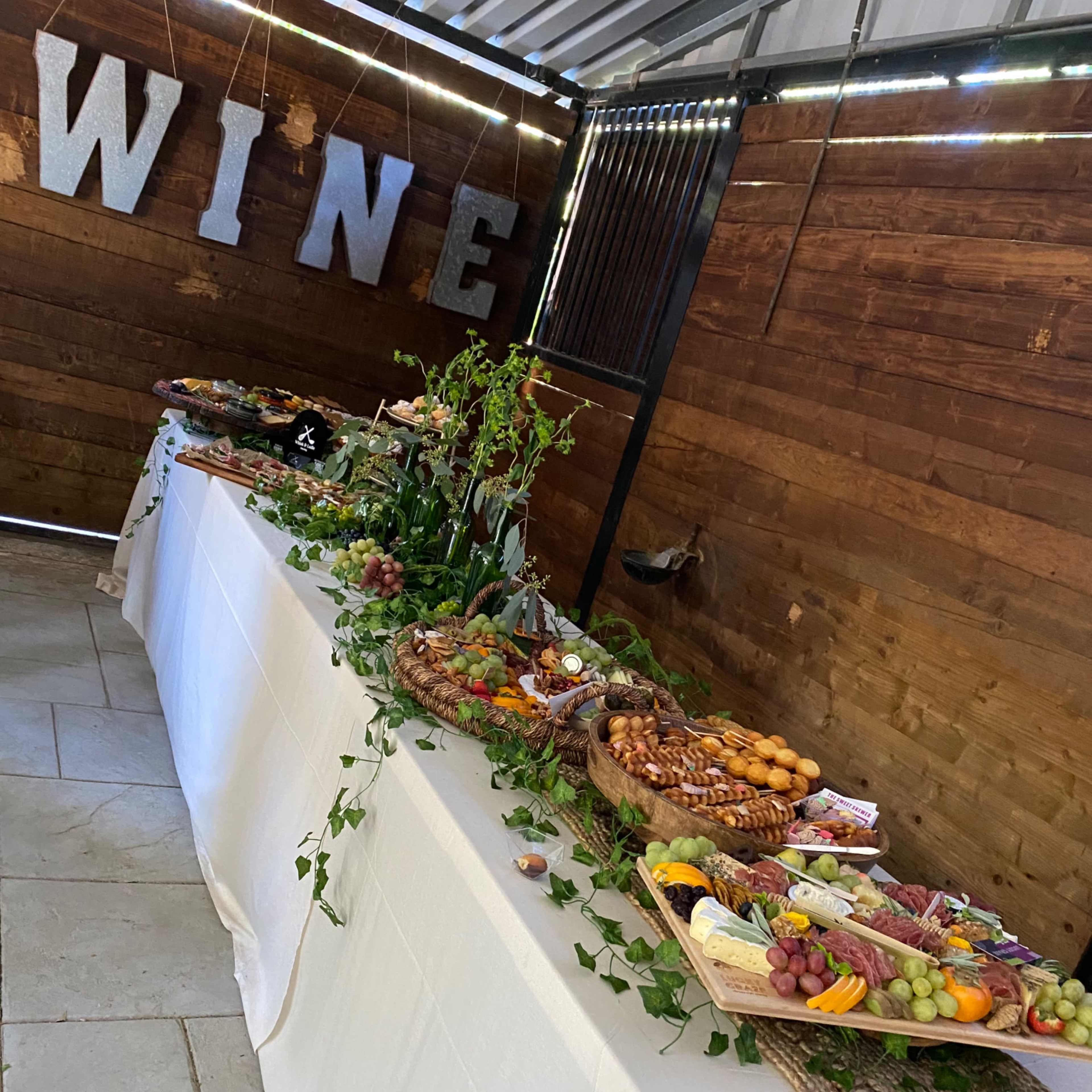 A long table covered with a white tablecloth features an assortment of fruits, cheeses, and meats, with decorative elements and the word "WINE" displayed in large letters on the wall behind it.
