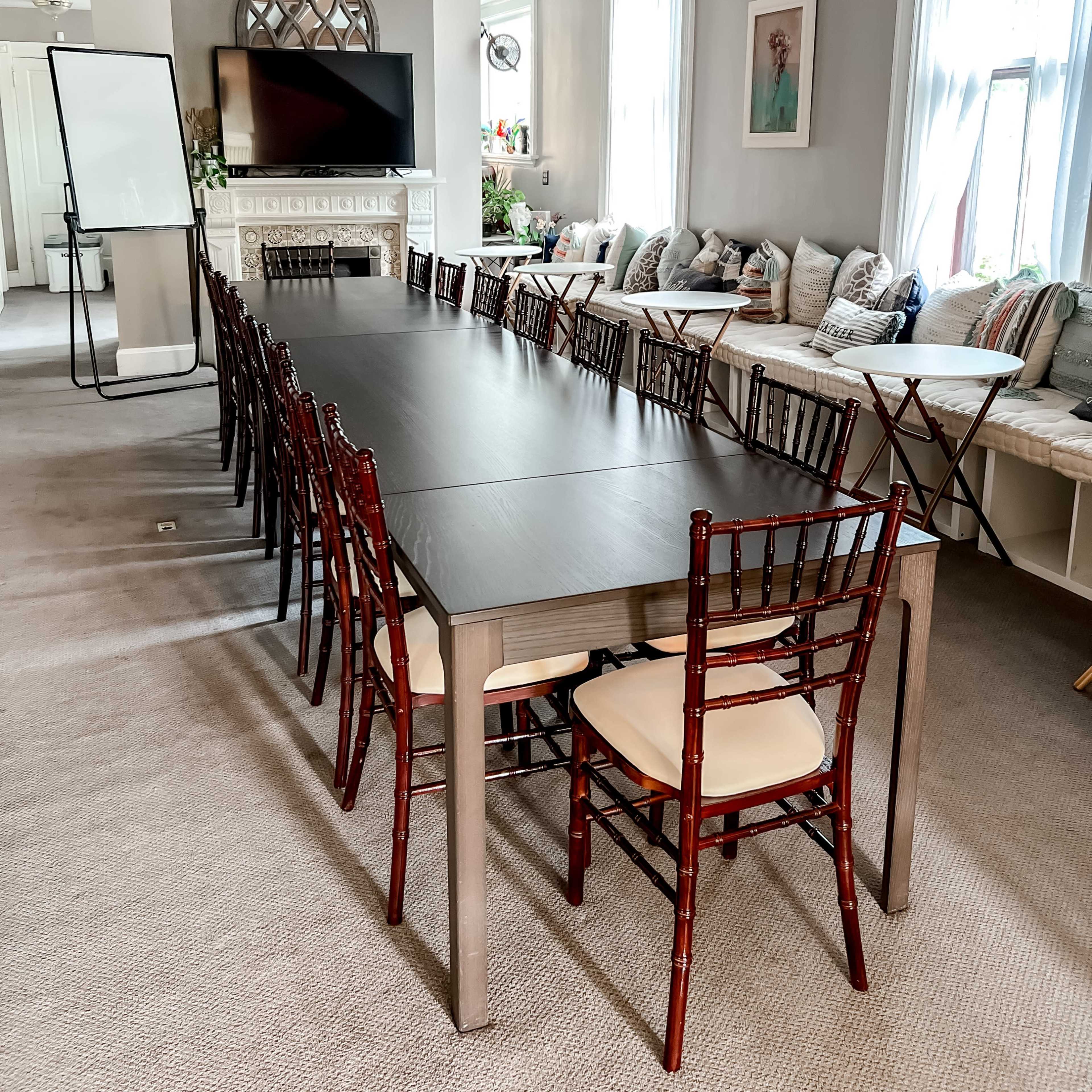 A long conference table is set up in a well-lit room with a fireplace, surrounded by chairs and featuring large windows with decorative cushions nearby.