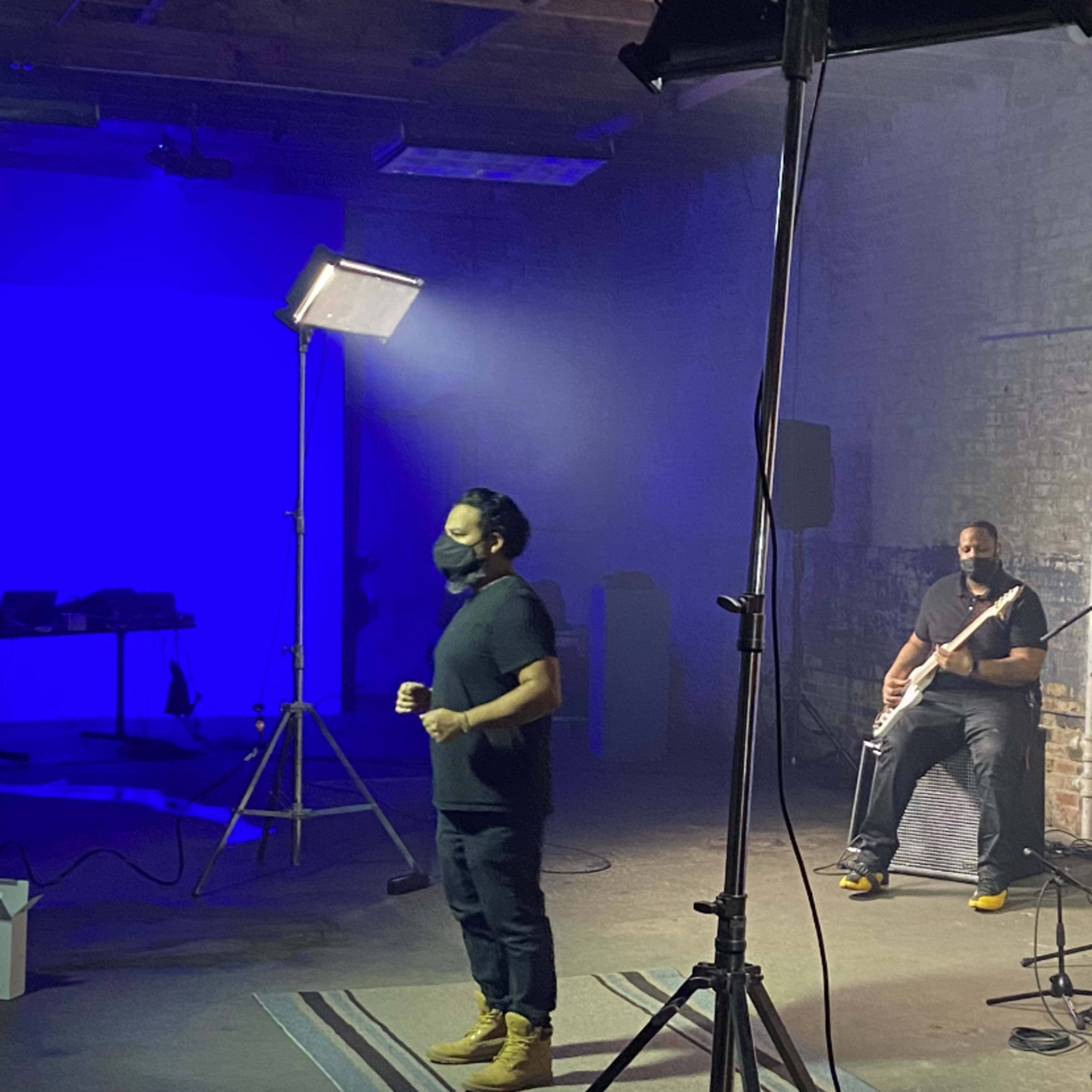 A band performs in a dimly lit warehouse with a blue backdrop and studio lighting.