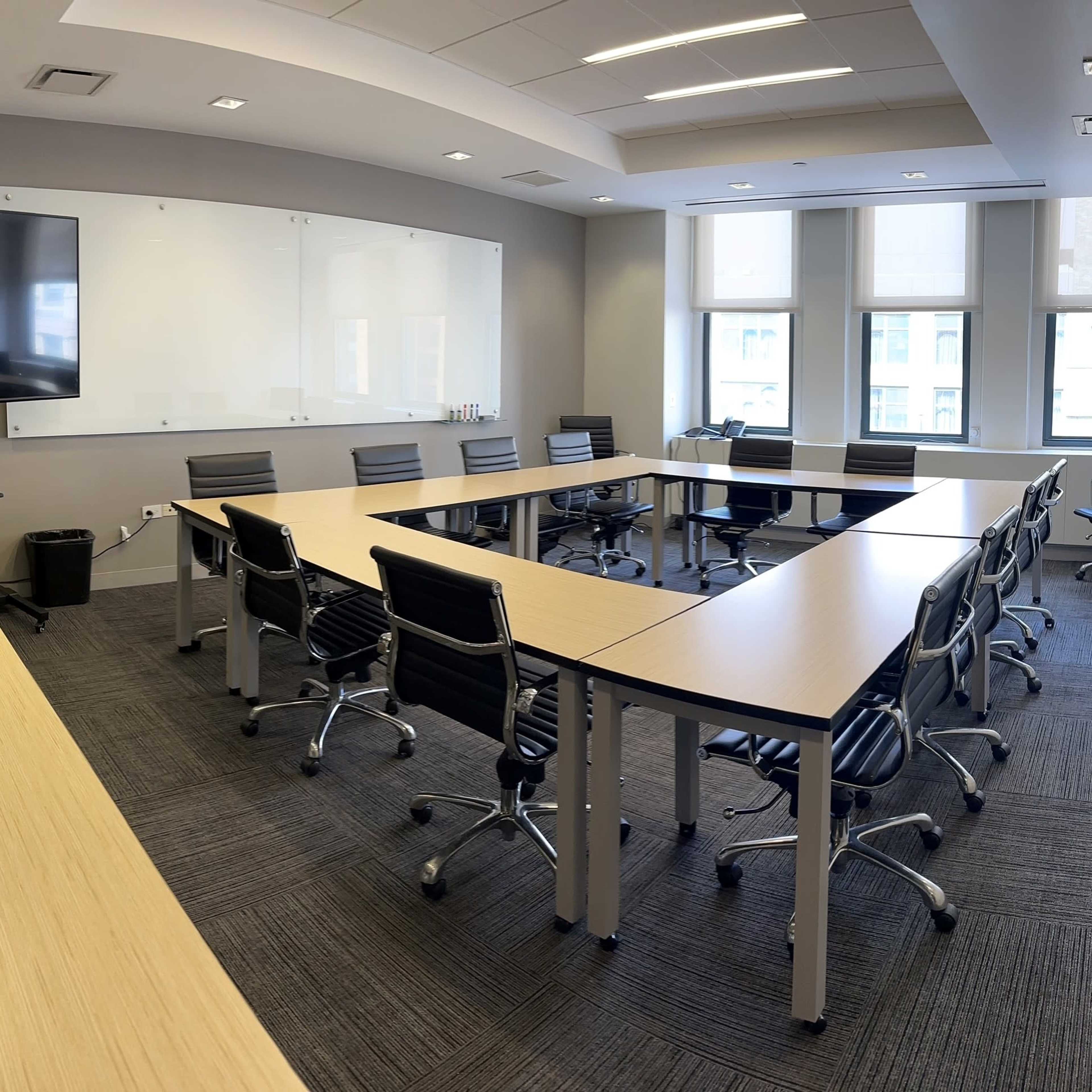 A modern conference room features a large rectangular table surrounded by ergonomic chairs, with a wall-mounted screen and windows offering natural light.