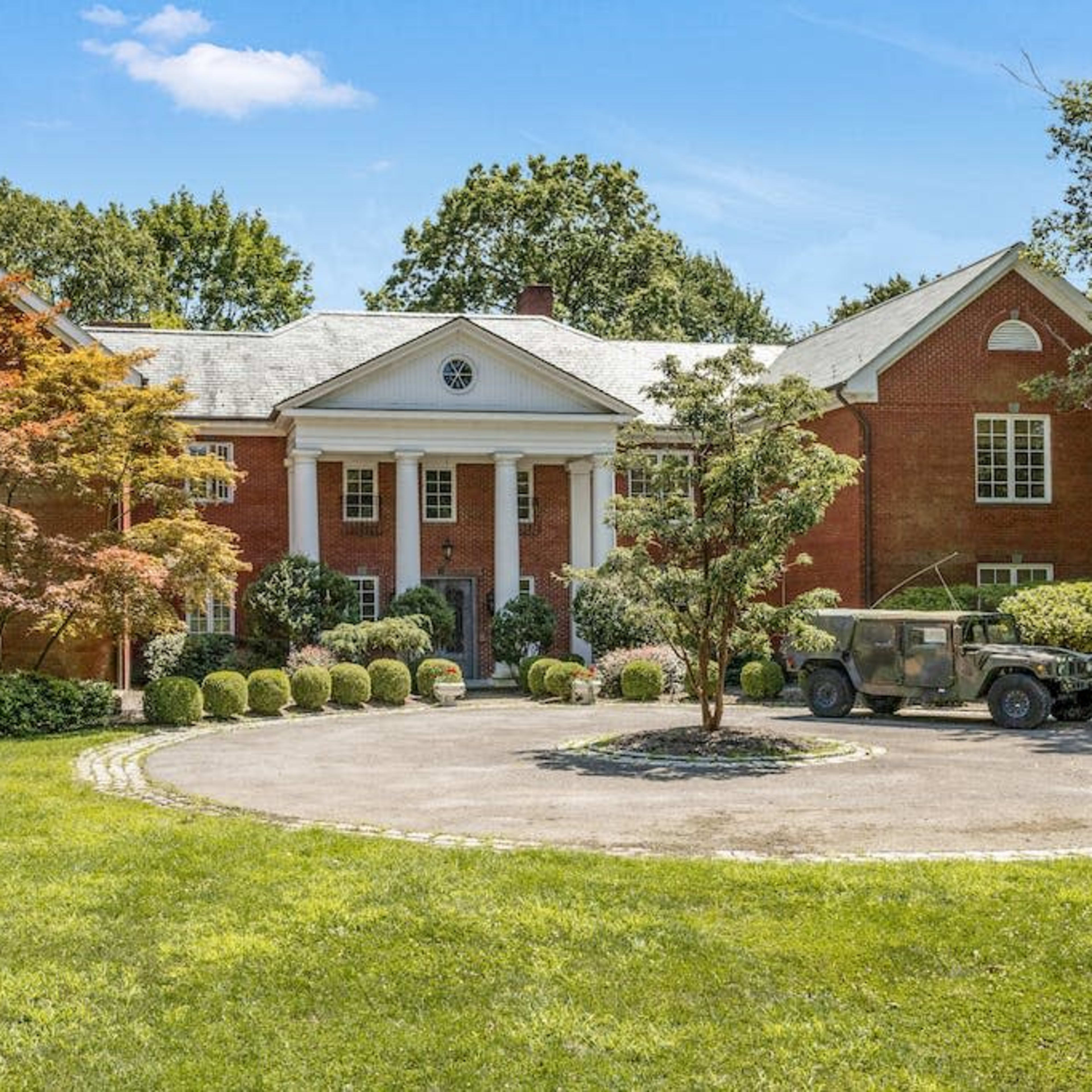 A large red brick mansion with a circular driveway, surrounded by landscaped greenery and a military vehicle parked in front.
