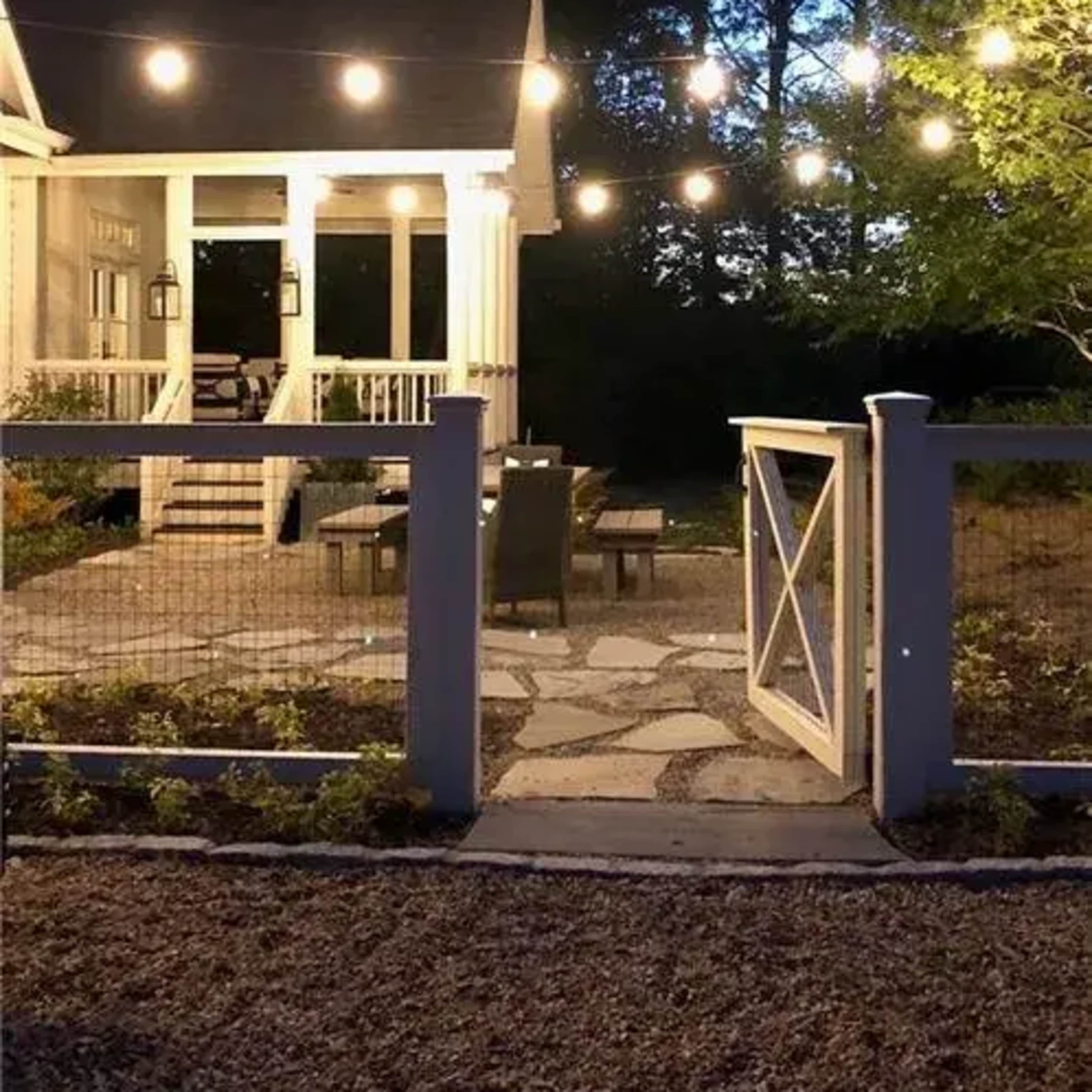A backyard area with string lights illuminating a seating arrangement on a stone patio, framed by a white gate and surrounded by trees at dusk.