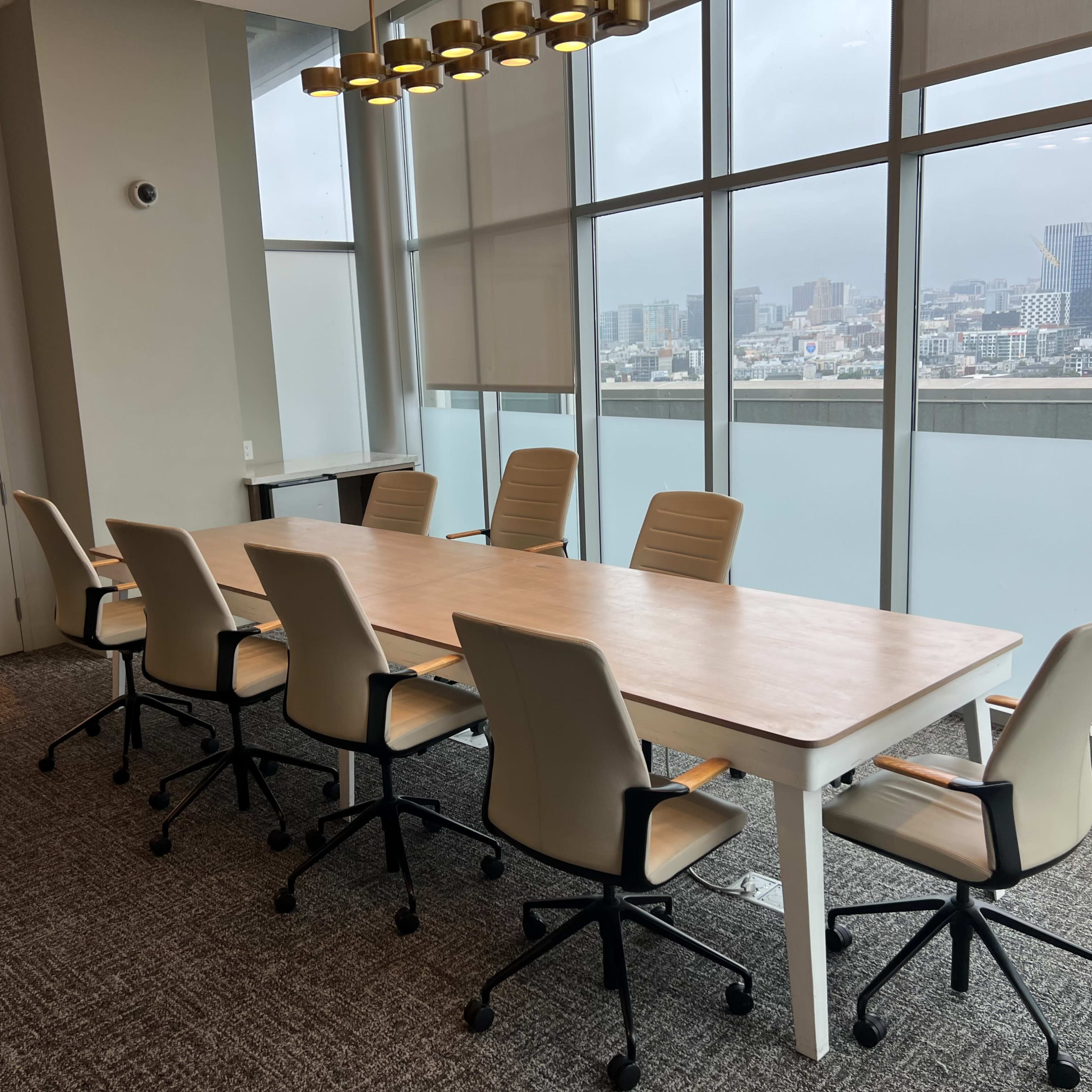 A large conference room features a long table surrounded by six rolling chairs, with floor-to-ceiling windows overlooking a city skyline.