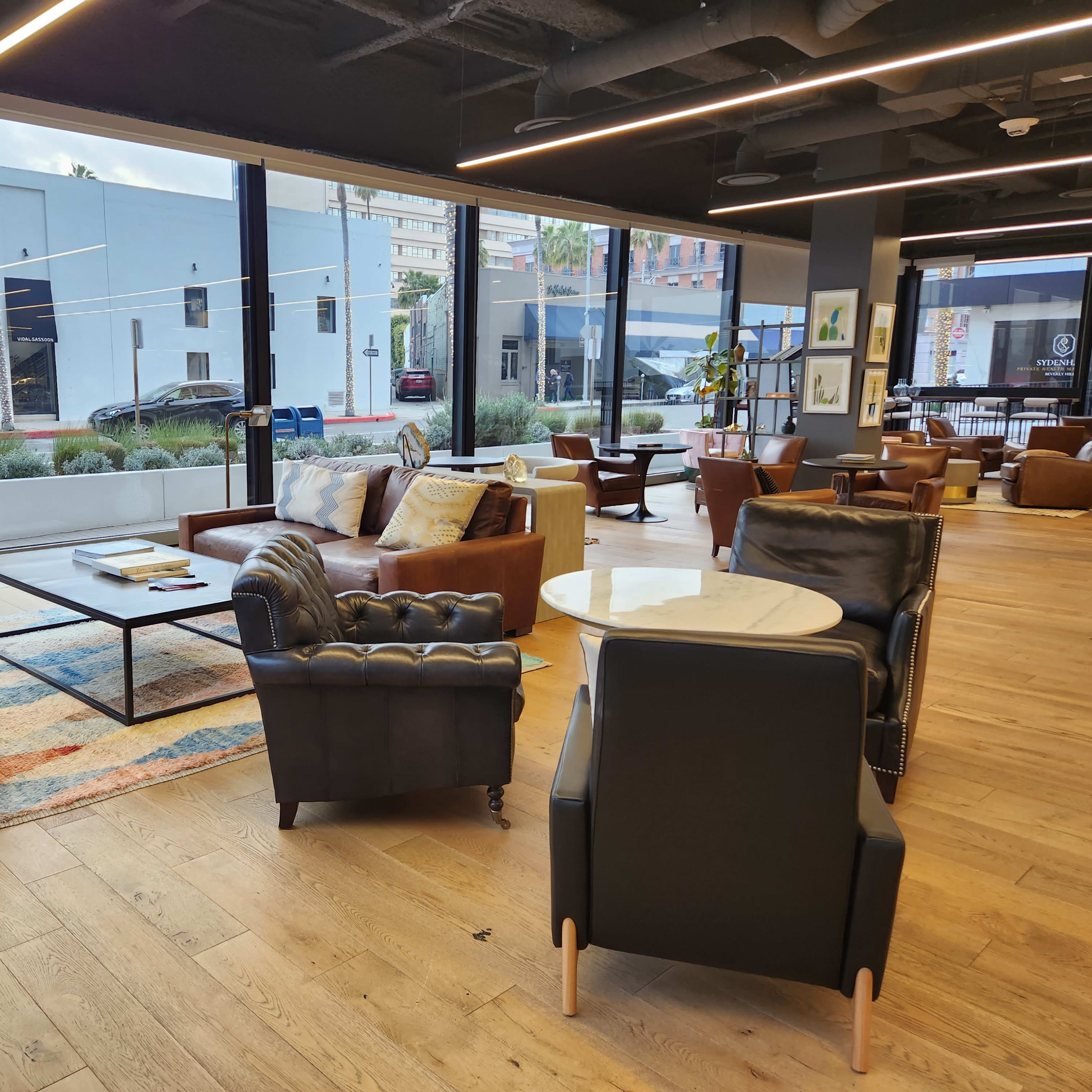 A cozy lounge area with large windows, featuring leather sofas, wooden floors, and a mix of tables and chairs.