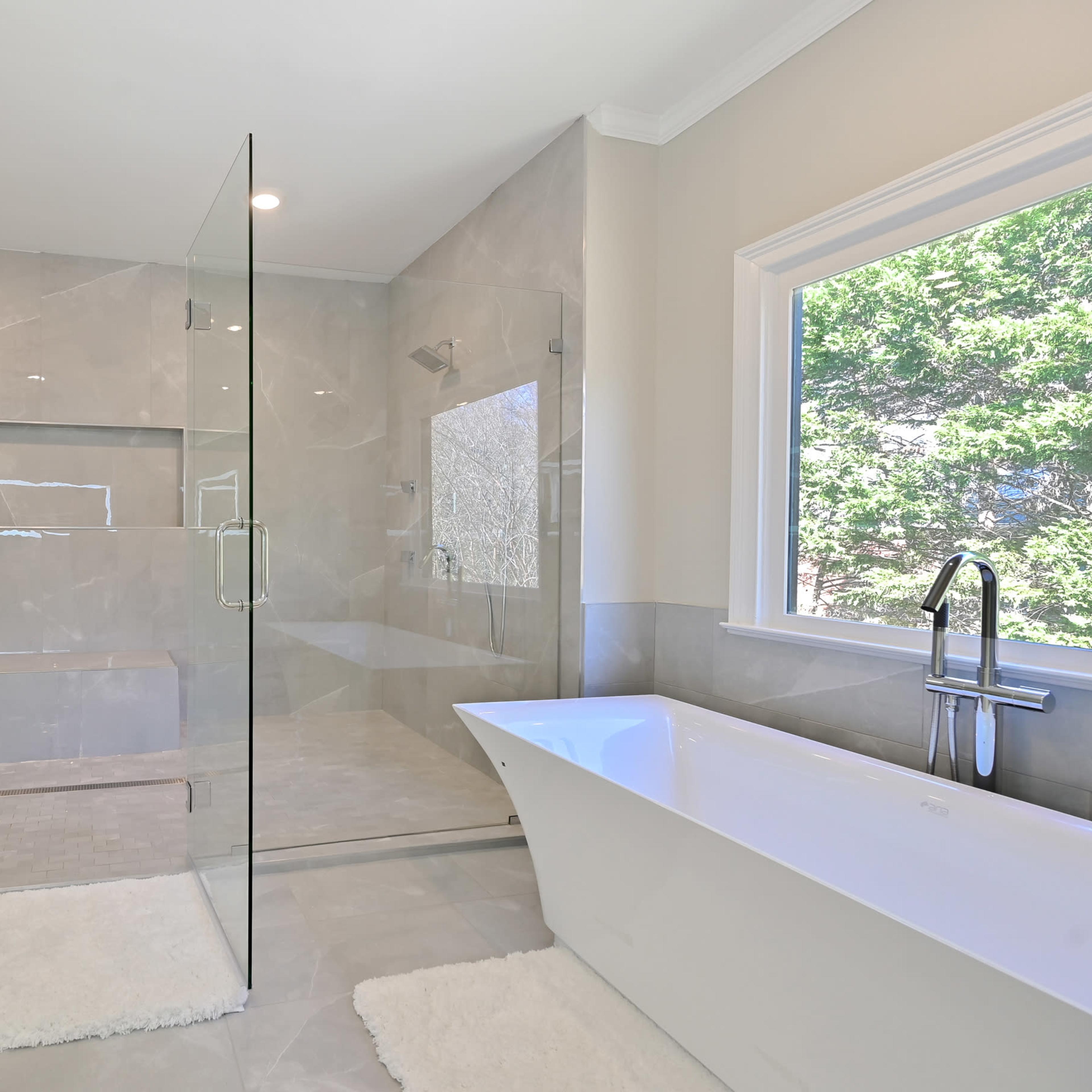 A modern bathroom featuring a glass shower enclosure, a freestanding bathtub, and light gray tile walls.