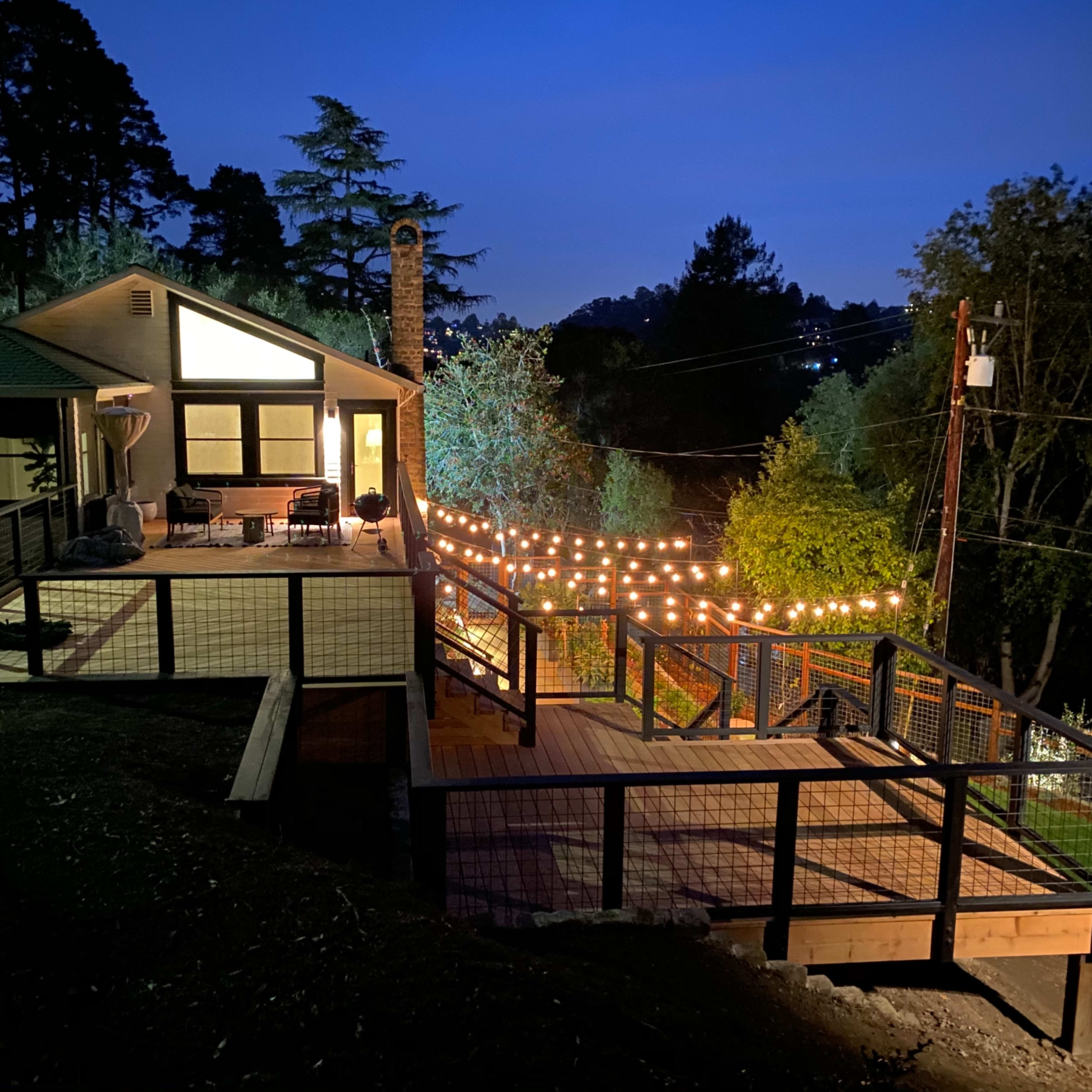 A modern house with a large deck and string lights is illuminated at night, surrounded by trees and power lines.