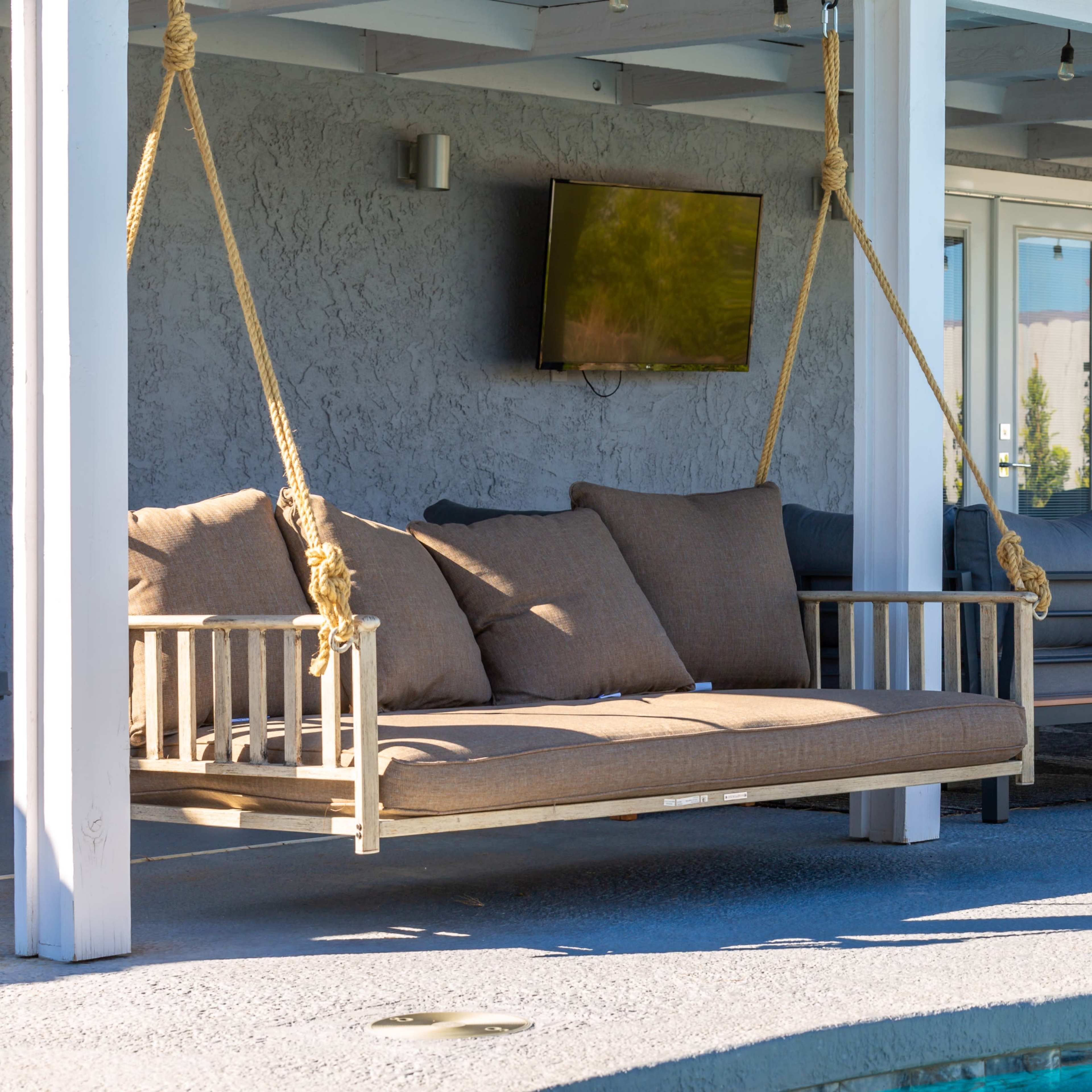 A wooden swing seat with cushions, suspended on ropes beneath a covered patio facing a pool.