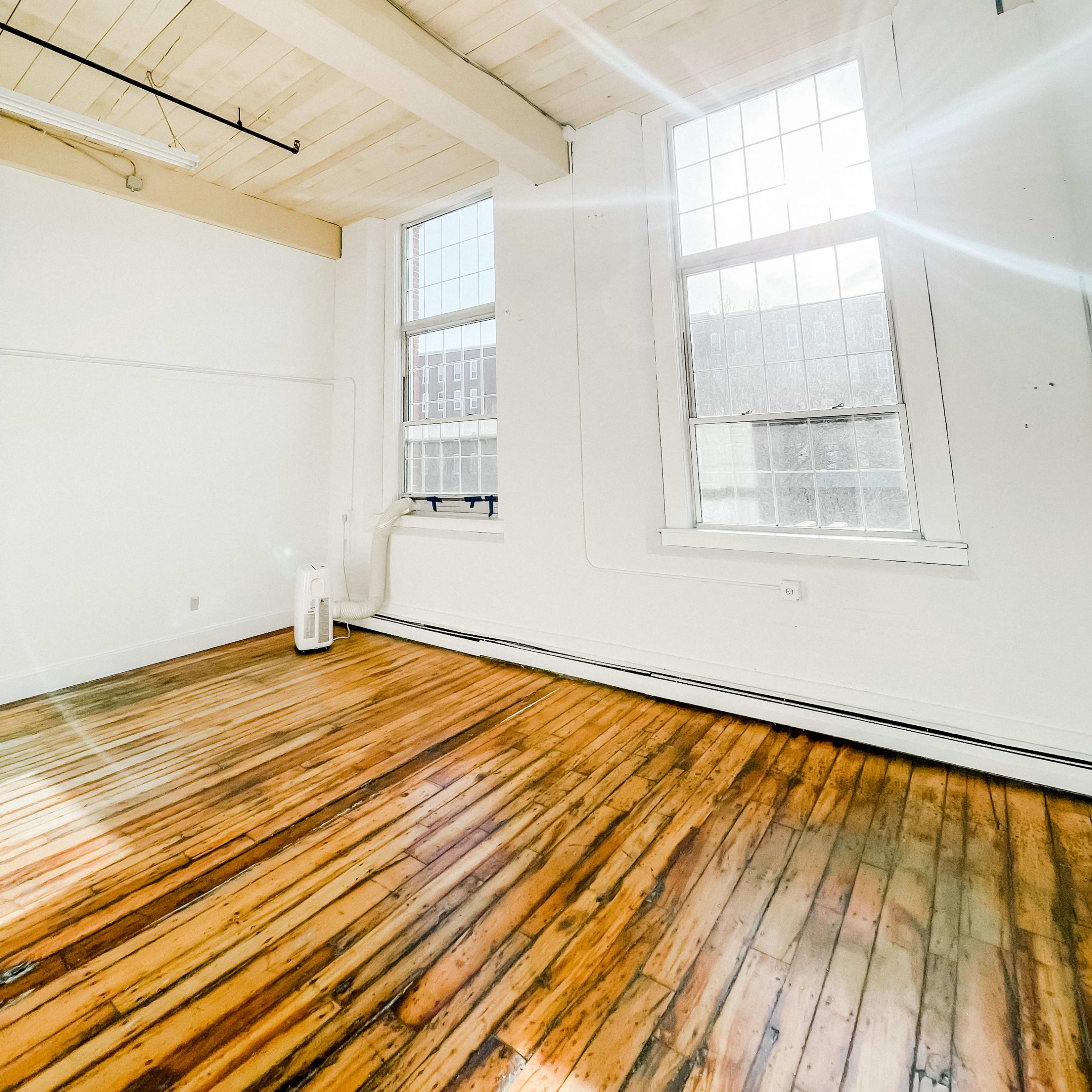 An empty room with large windows allowing natural light to illuminate the polished wooden floor and white walls.