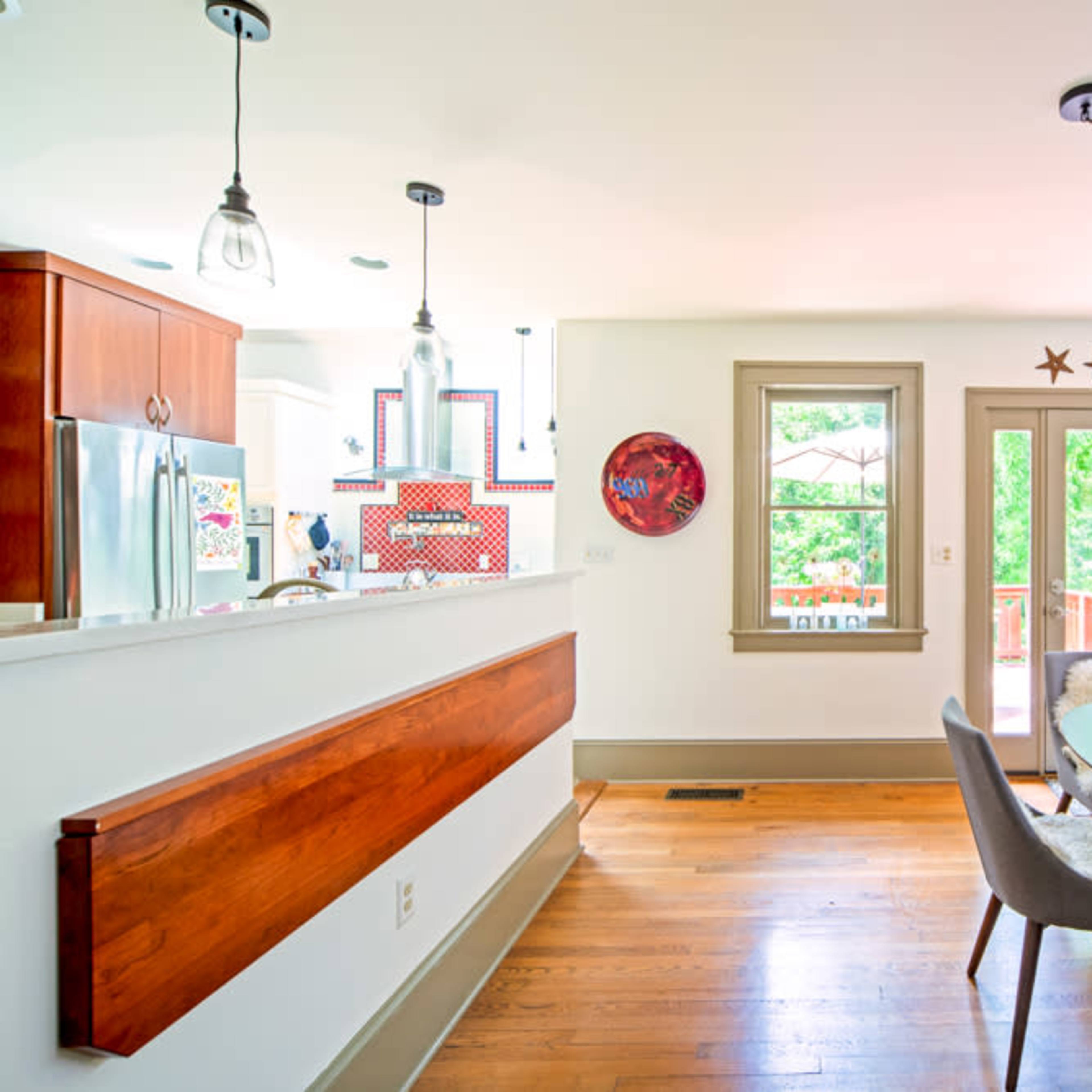 A modern dining area features a round table surrounded by chairs, with a kitchen and large windows visible in the background.
