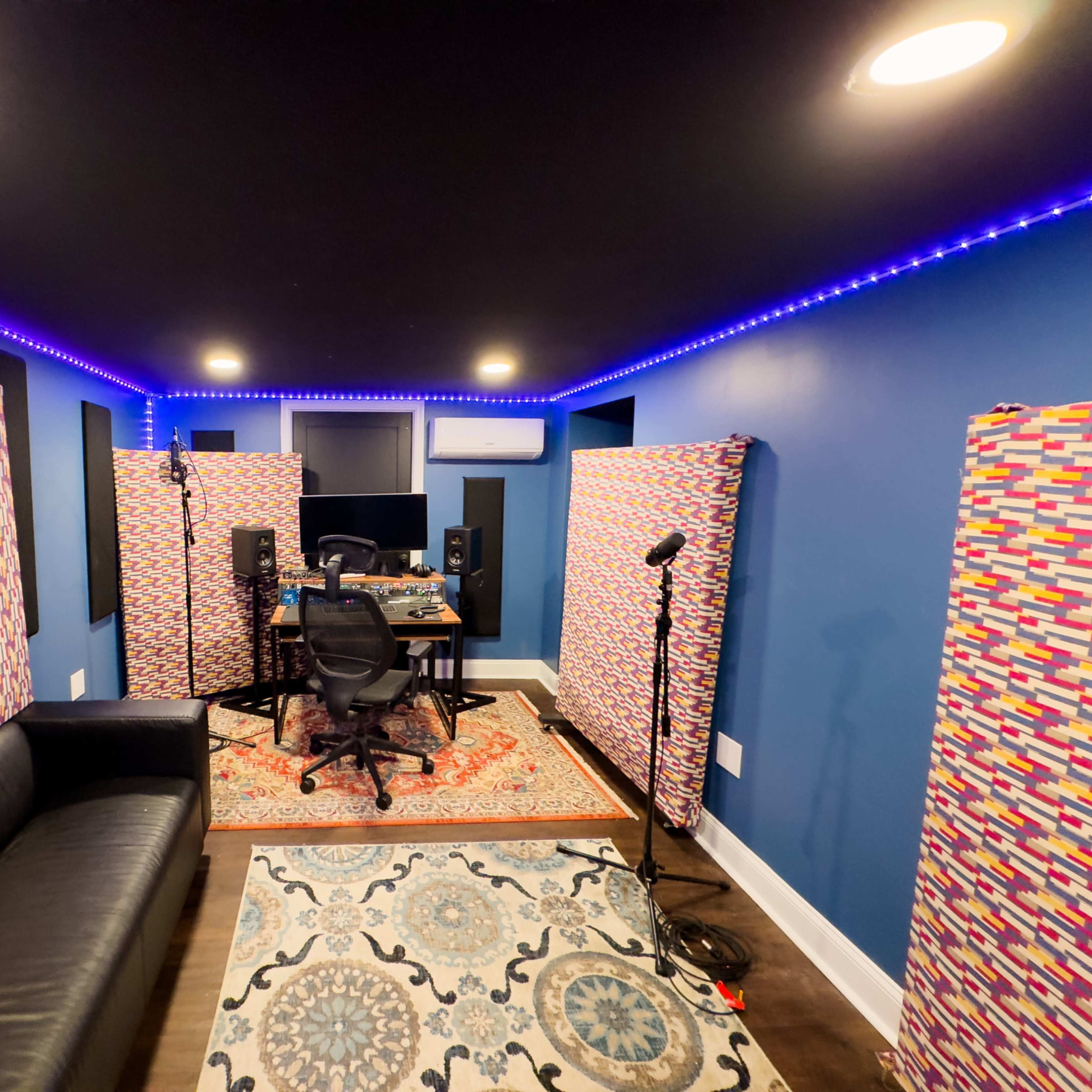 A music recording studio with soundproofing panels, microphones, and a desk equipped with audio equipment, illuminated by LED lights along the walls.