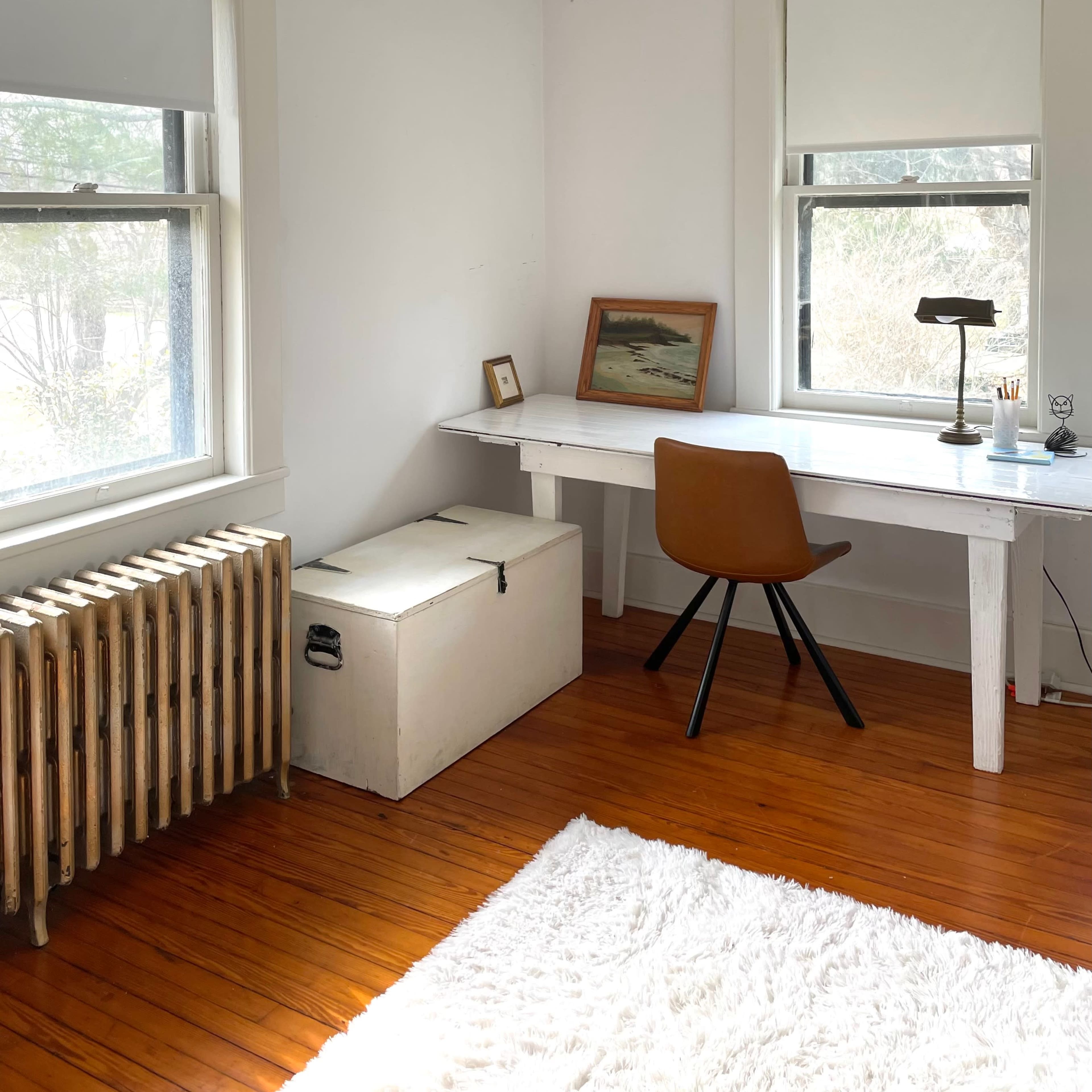 A bright room with a white desk, a brown chair, a vintage trunk, and a radiator near the windows.