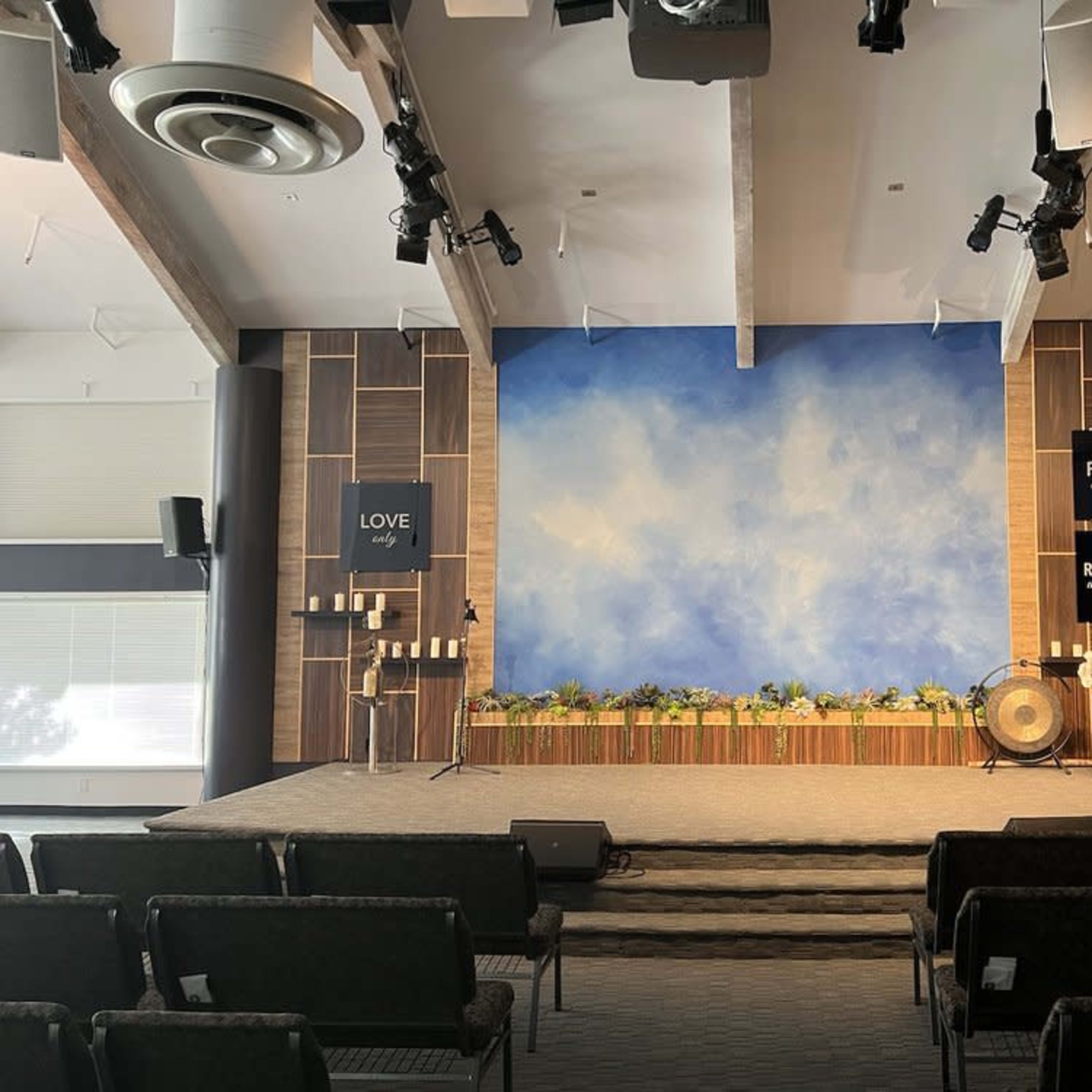A church stage with a blue sky backdrop, candles, and decorative plants, along with signs that read "LOVE," "FORGIVE," and "REMEMBER."