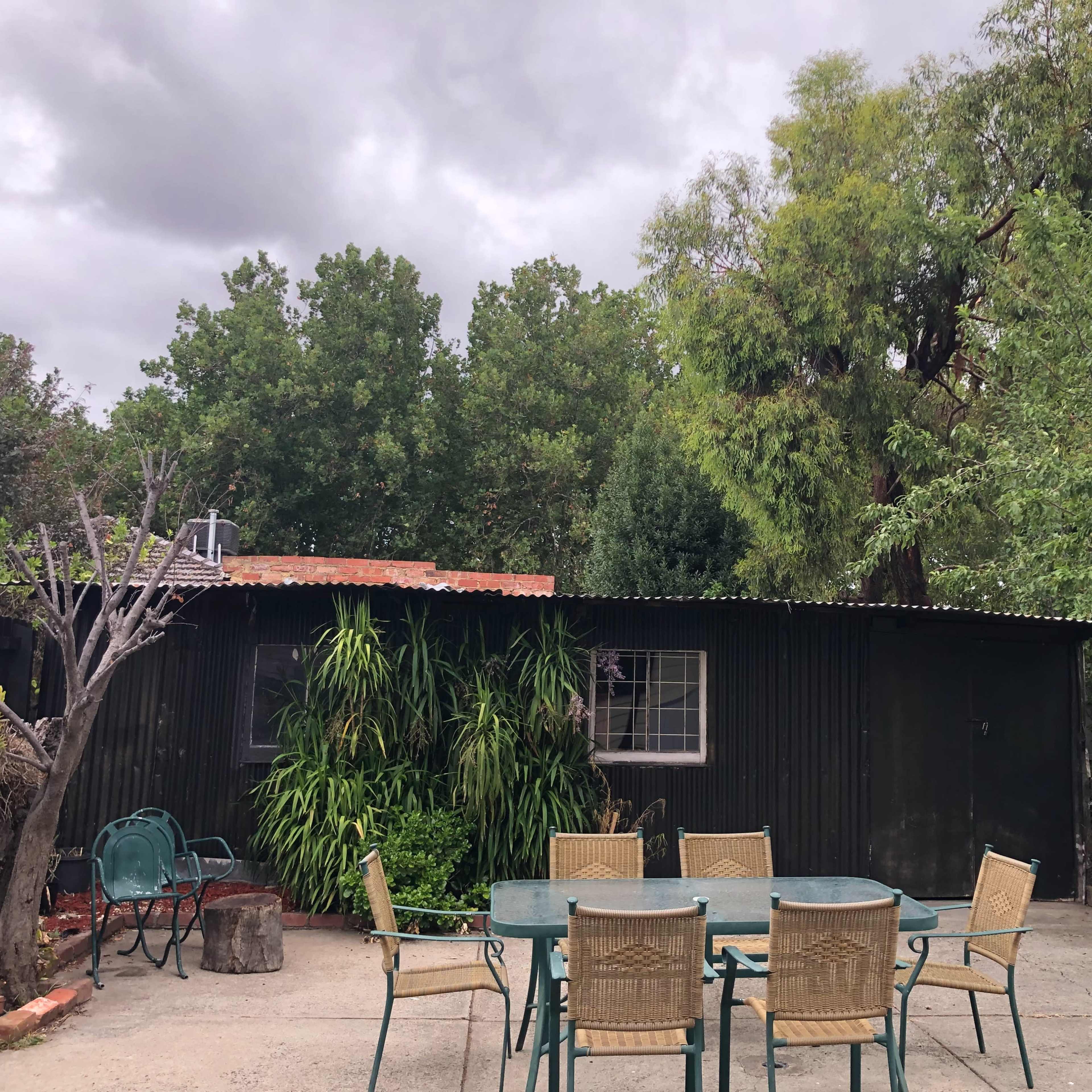 A small, dark green shed is set against a backdrop of trees, with a metal table and chairs in the foreground.
