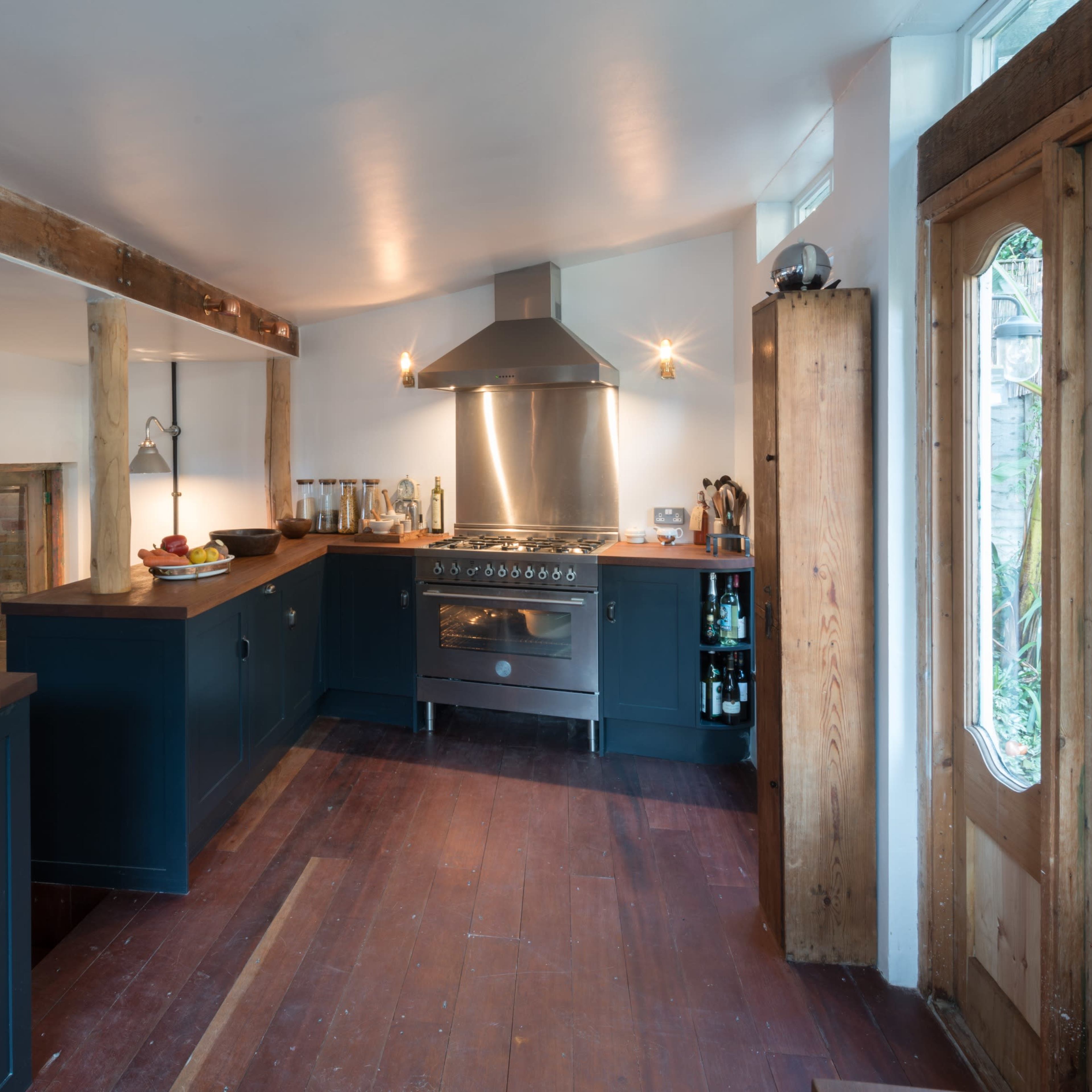 A modern kitchen with dark cabinets, a stainless steel stove, wooden beams, and a front door featuring stained glass.