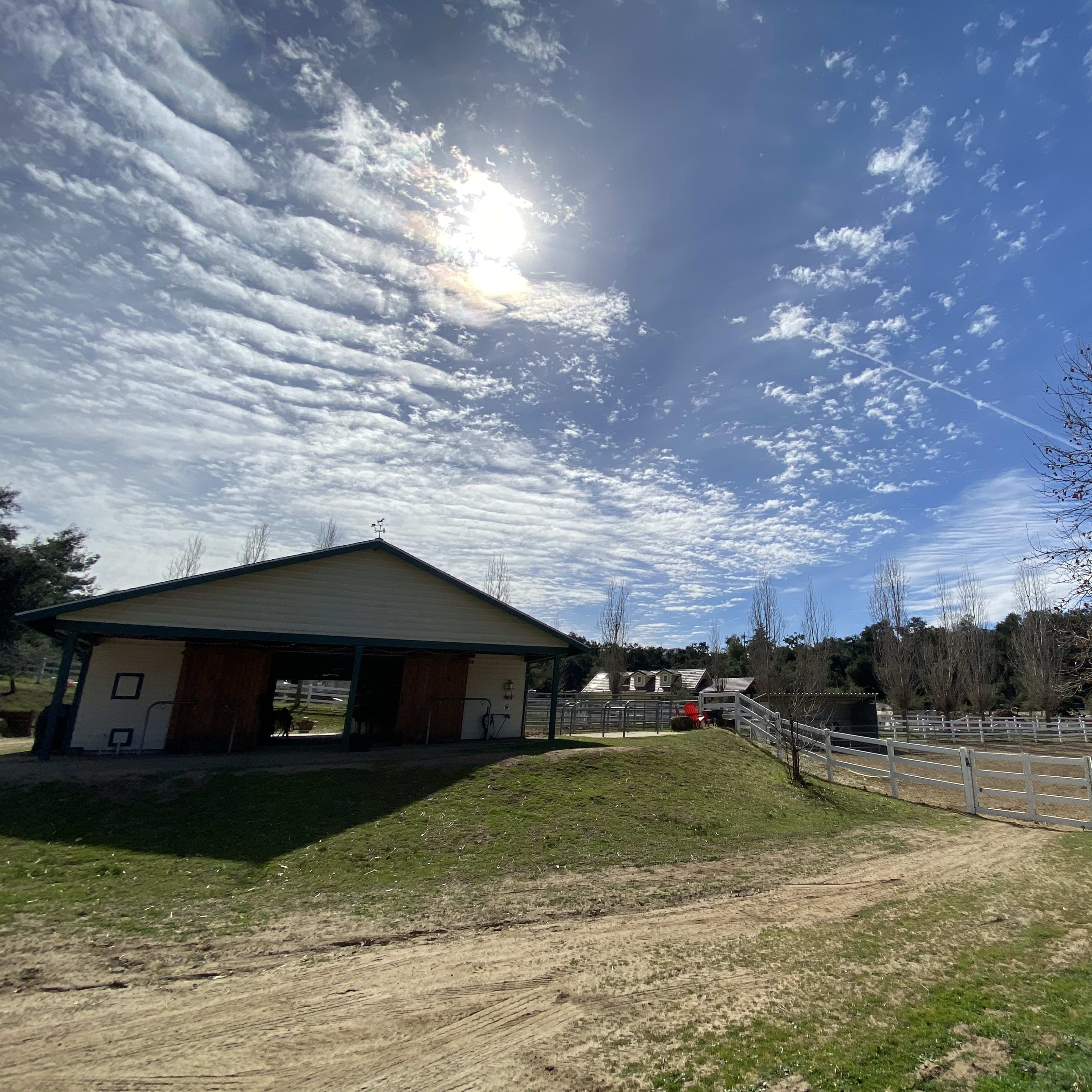 A barn sits beside a fenced horse paddock under a partly cloudy sky.