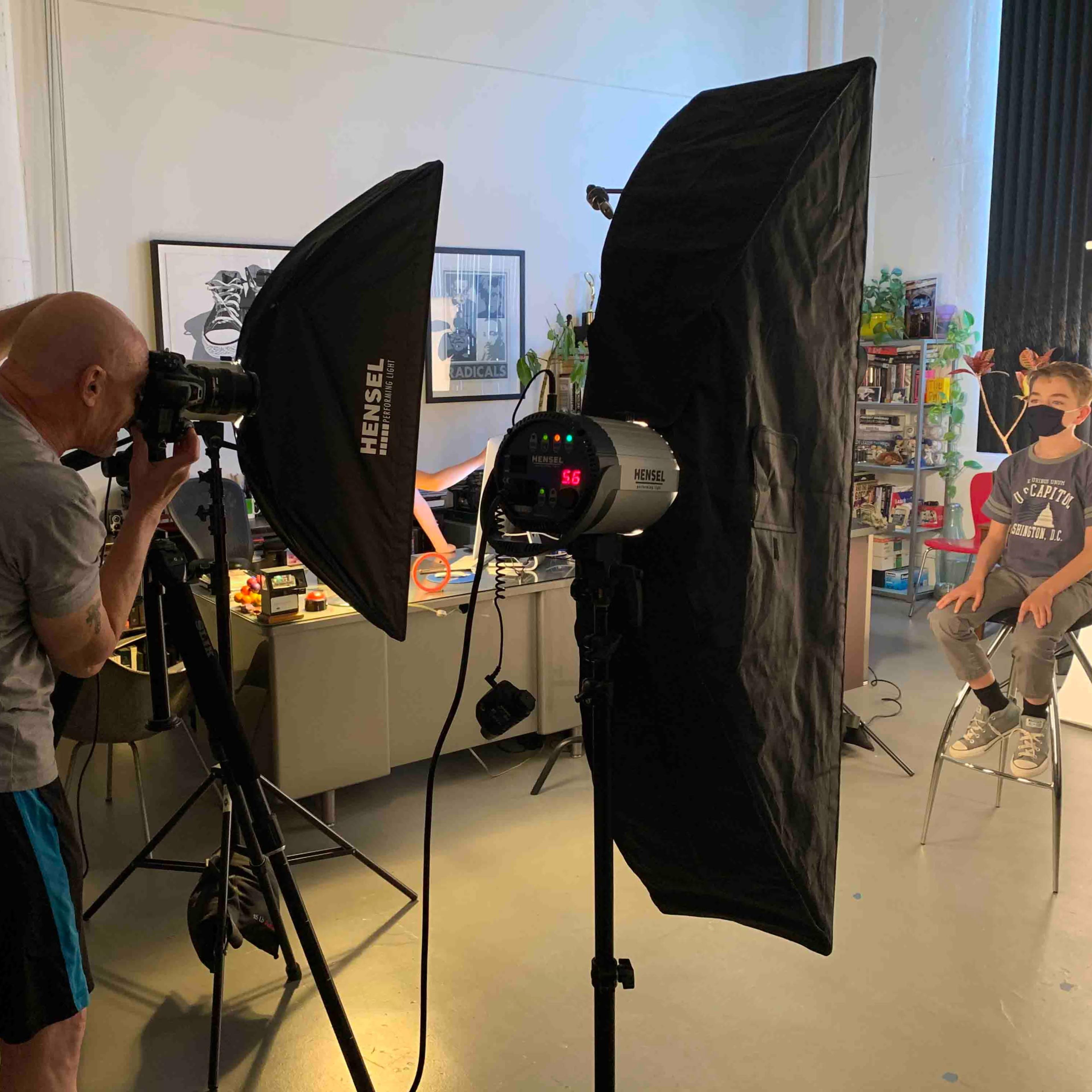 A photographer uses a camera to capture an image of a child sitting on a stool in a well-lit studio setup.