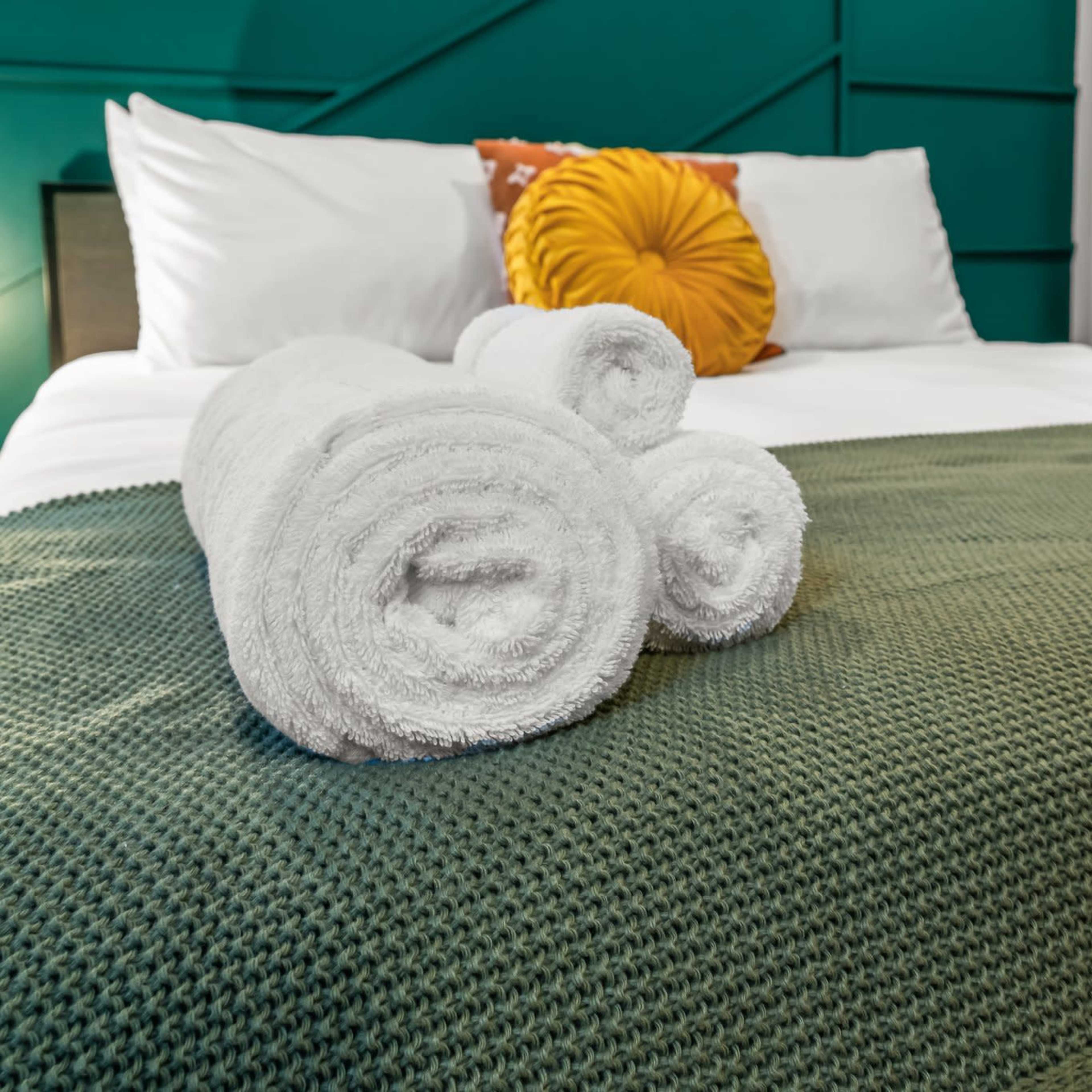 A neatly made bed with white towels rolled on a green blanket and a decorative yellow pillow.