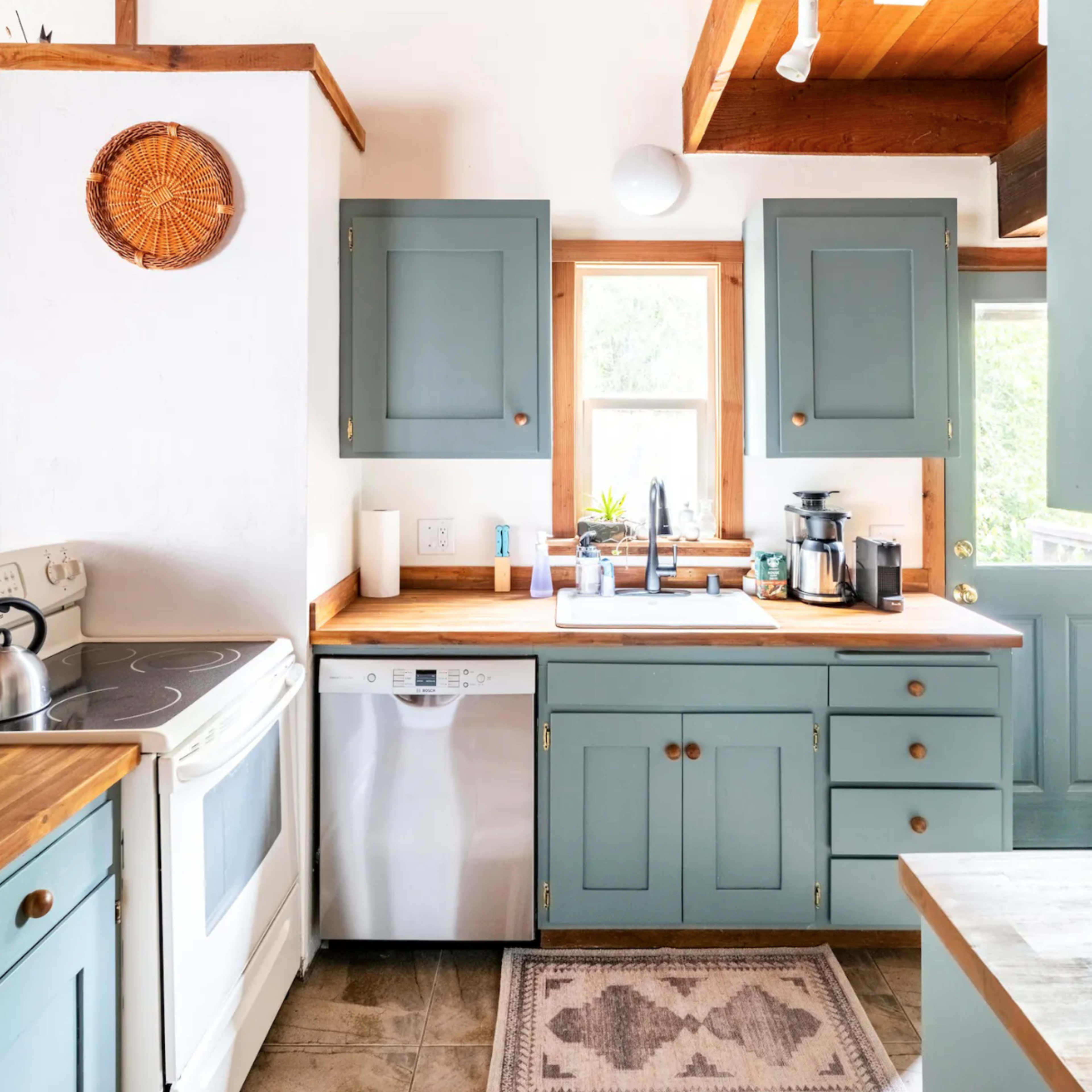A kitchen with pale blue cabinets, a white stove, a dishwasher, and a wooden countertop, featuring natural light from a nearby window.