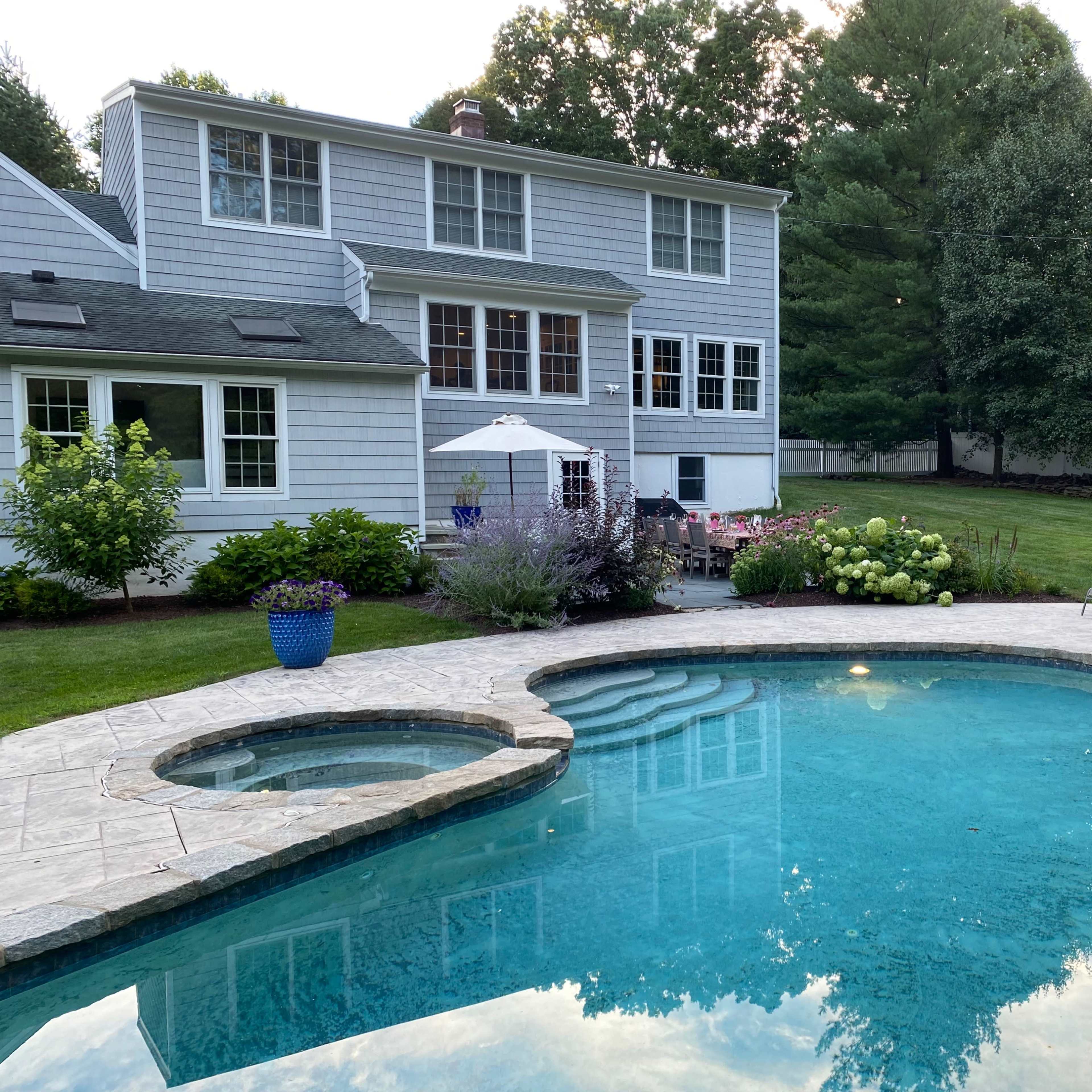 A backyard scene features a blue swimming pool surrounded by patio furniture, landscaping, and a two-story grey house in the background.