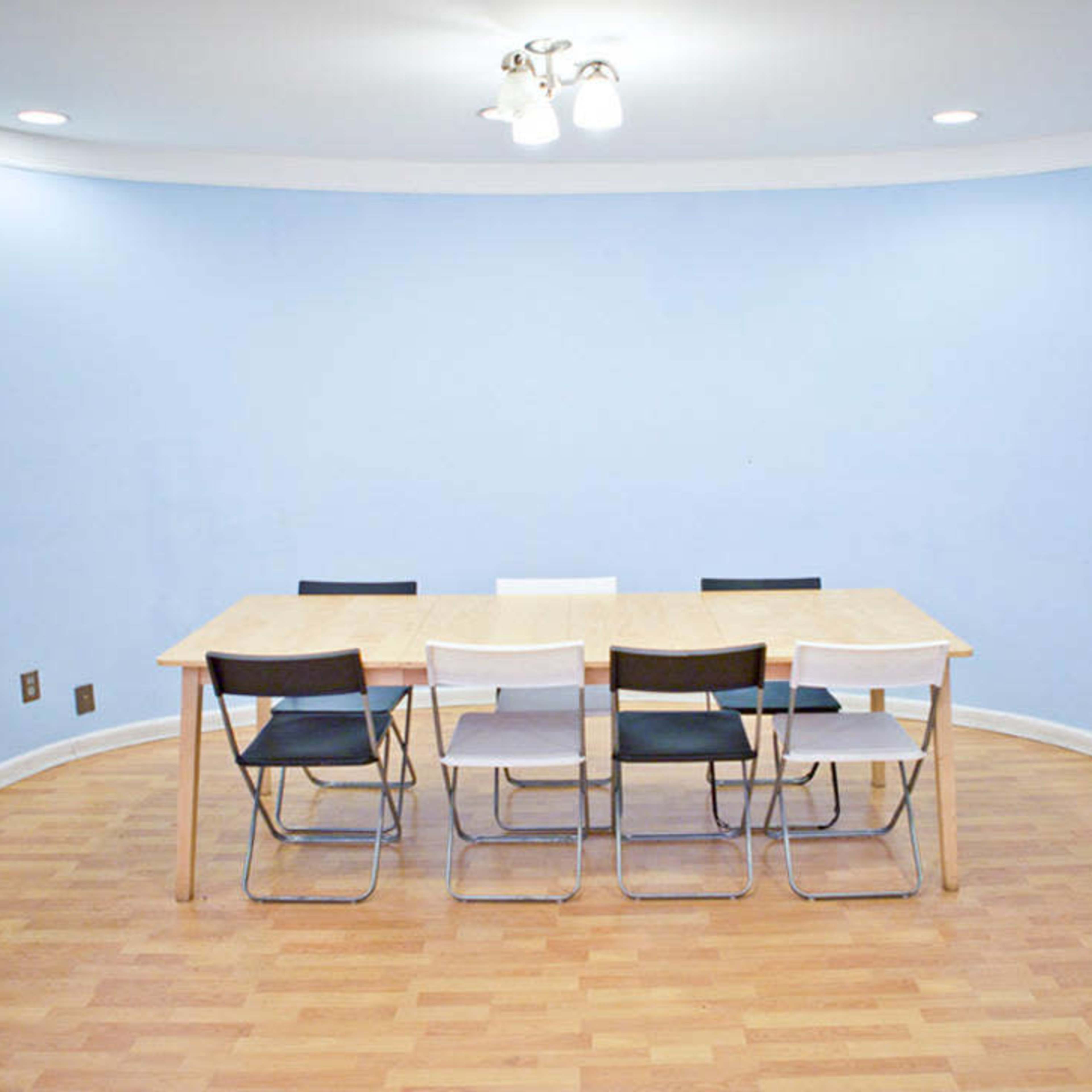 A wooden table with six folding chairs is positioned in a light blue room with curved walls and wooden flooring.