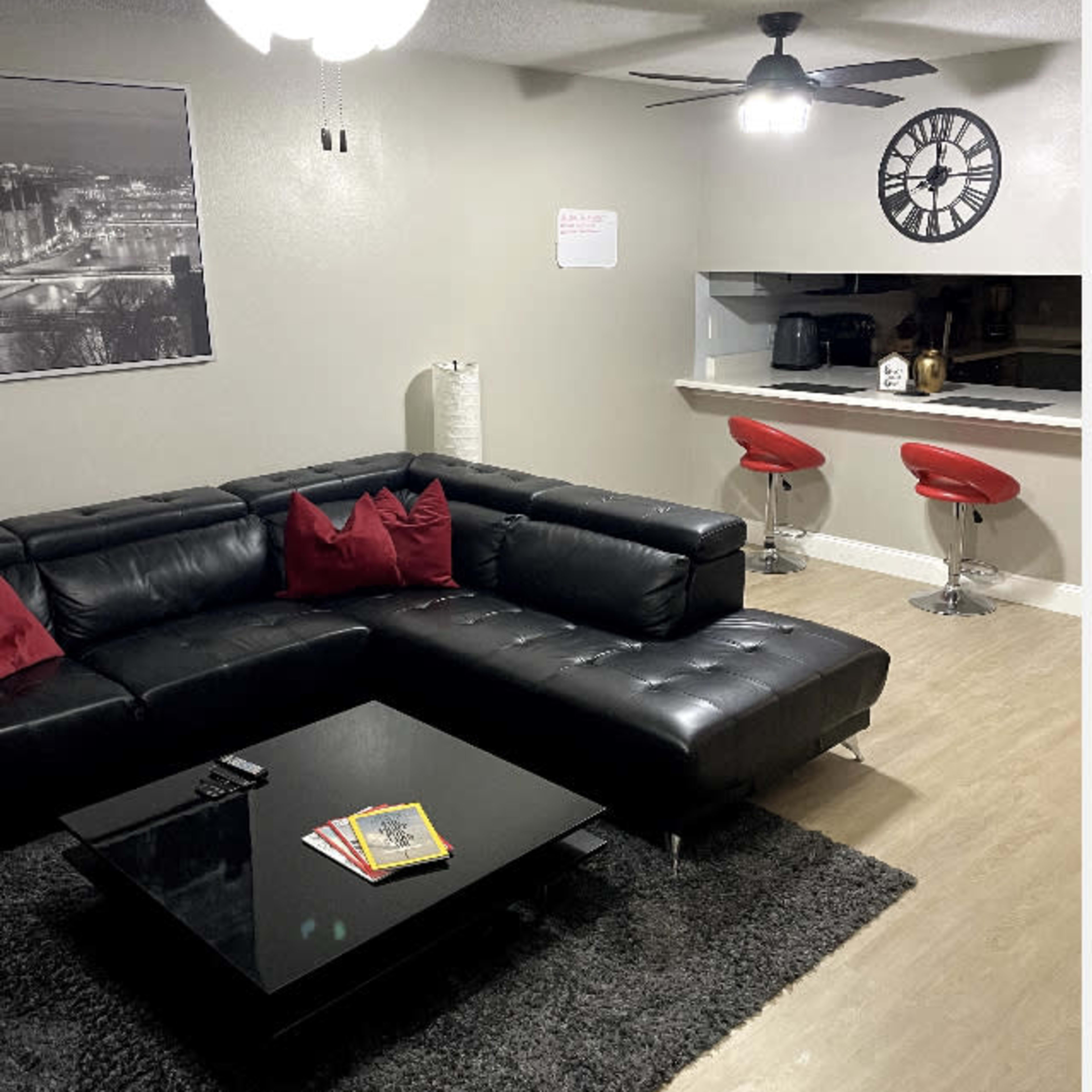A modern living room features a black sectional sofa with red cushions, a glass coffee table on a rug, and a bar area with red stools in the background.