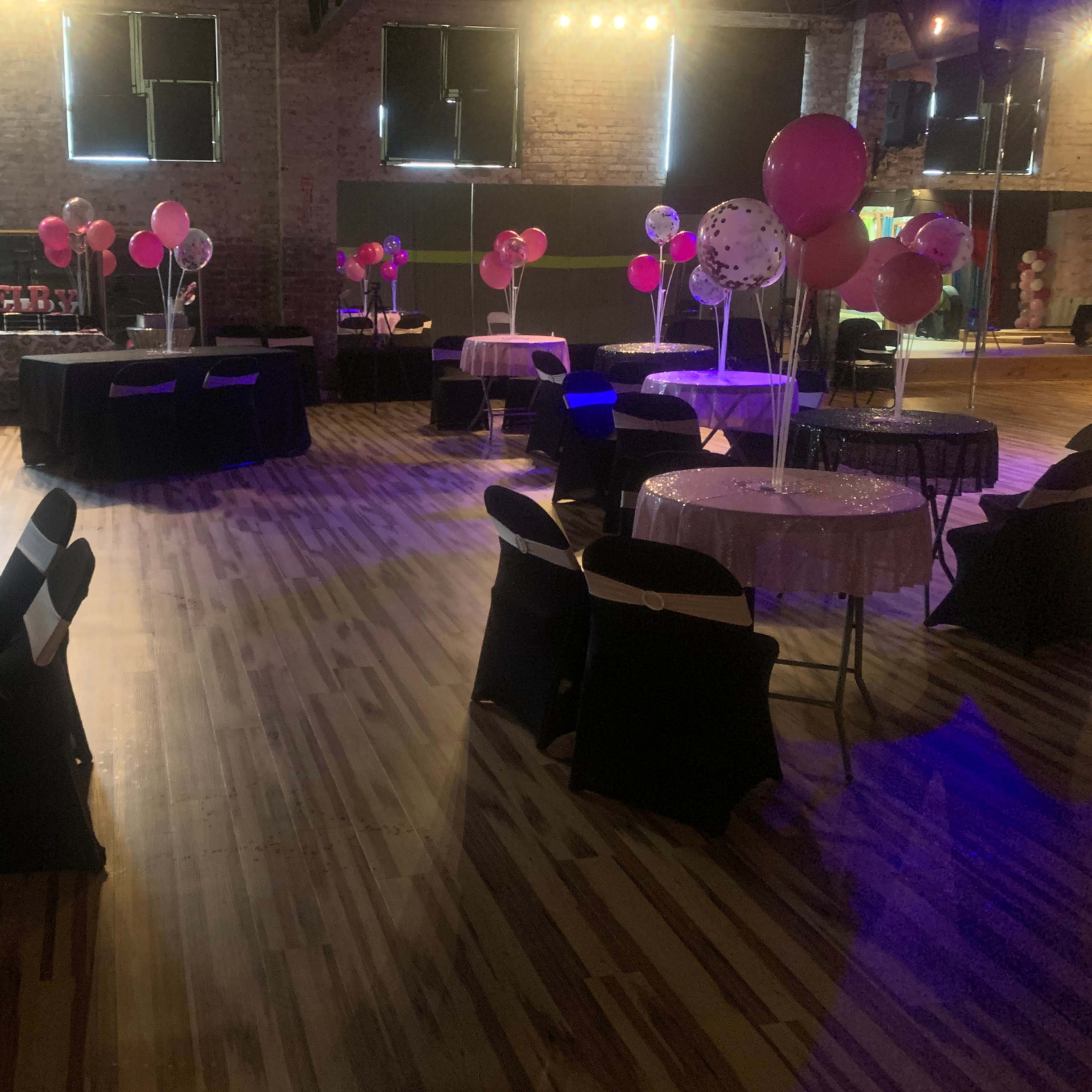 A spacious event venue features black and pink tables, balloons, and a polished wooden floor.