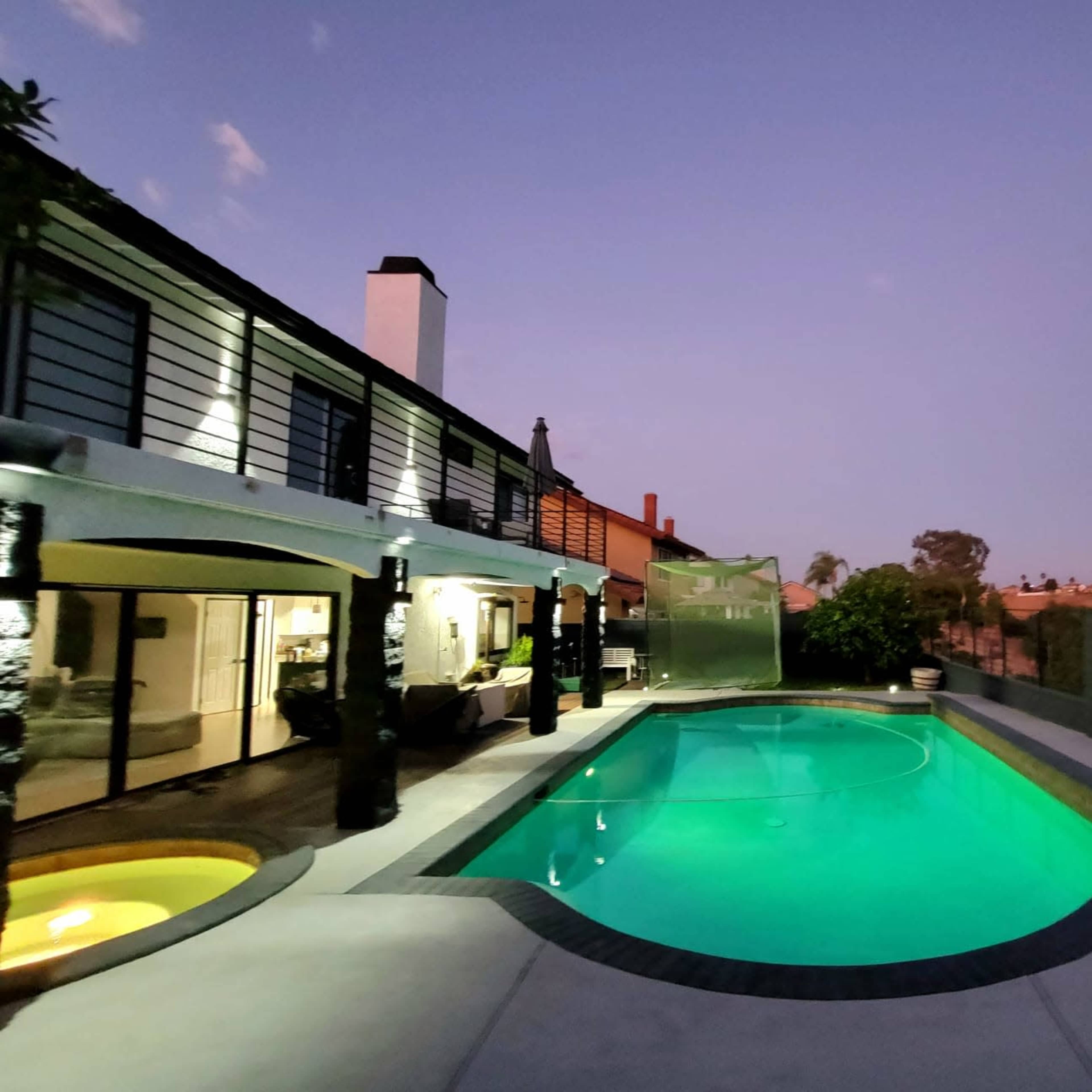 A modern house with a pool glows in the twilight, surrounded by landscaped grounds and hills in the background.