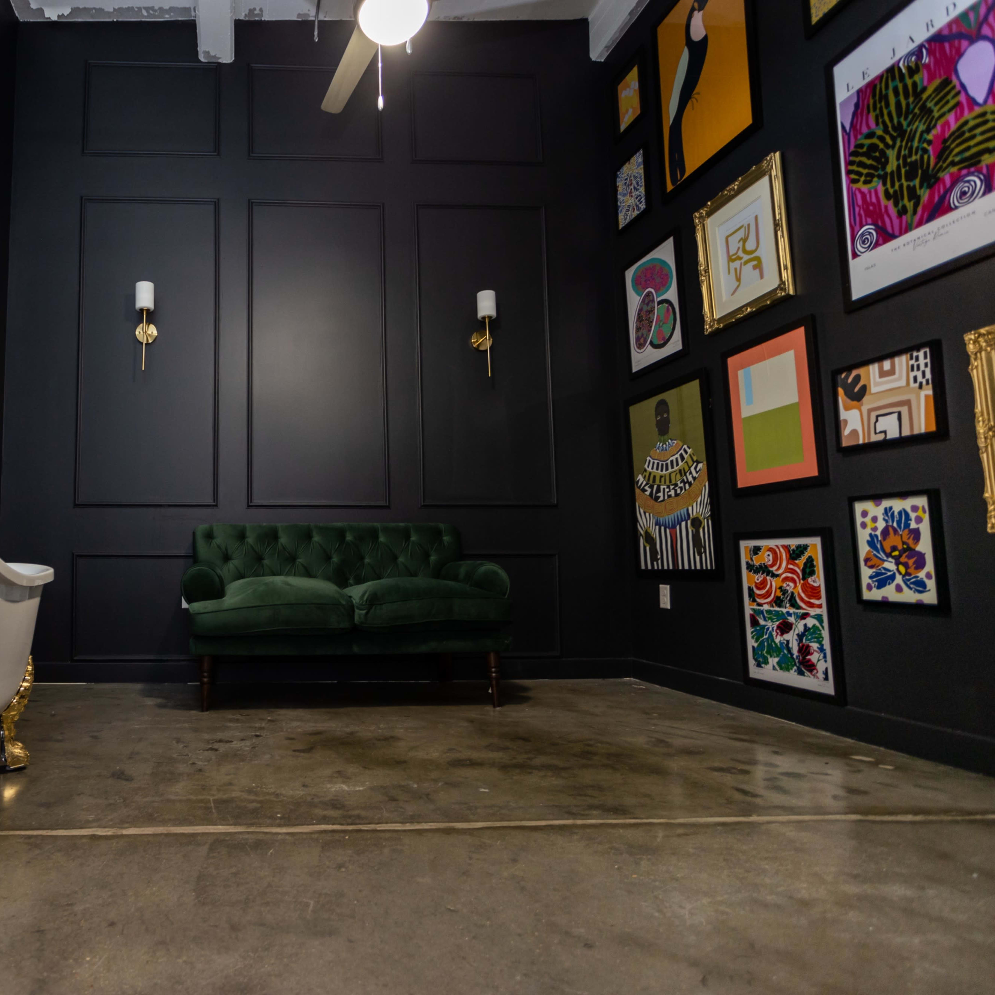 A dark room with a white clawfoot bathtub on one side and a green velvet couch opposite a wall adorned with various framed artworks.