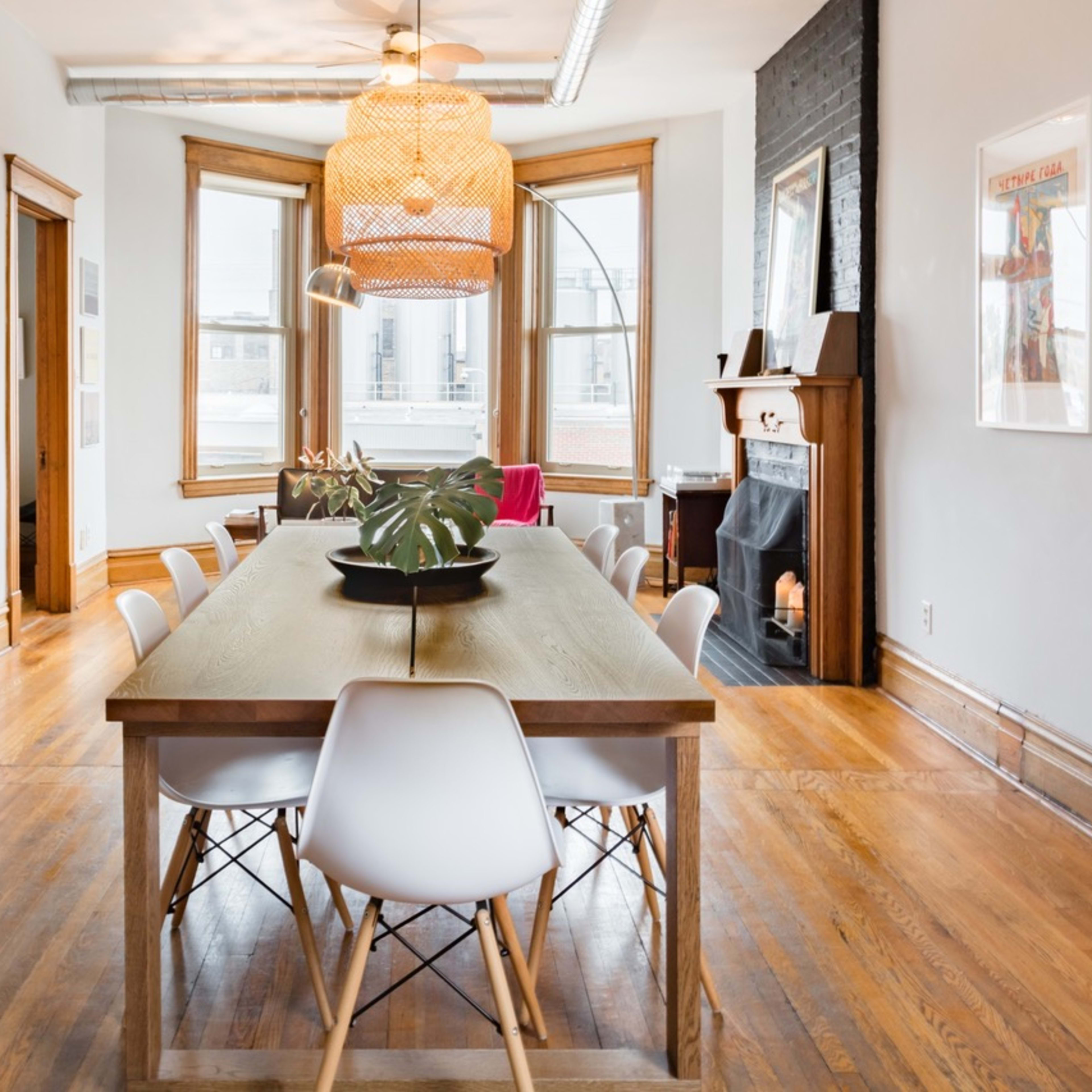 A dining area features a long wooden table surrounded by white chairs, with large windows and a decorative light fixture above.