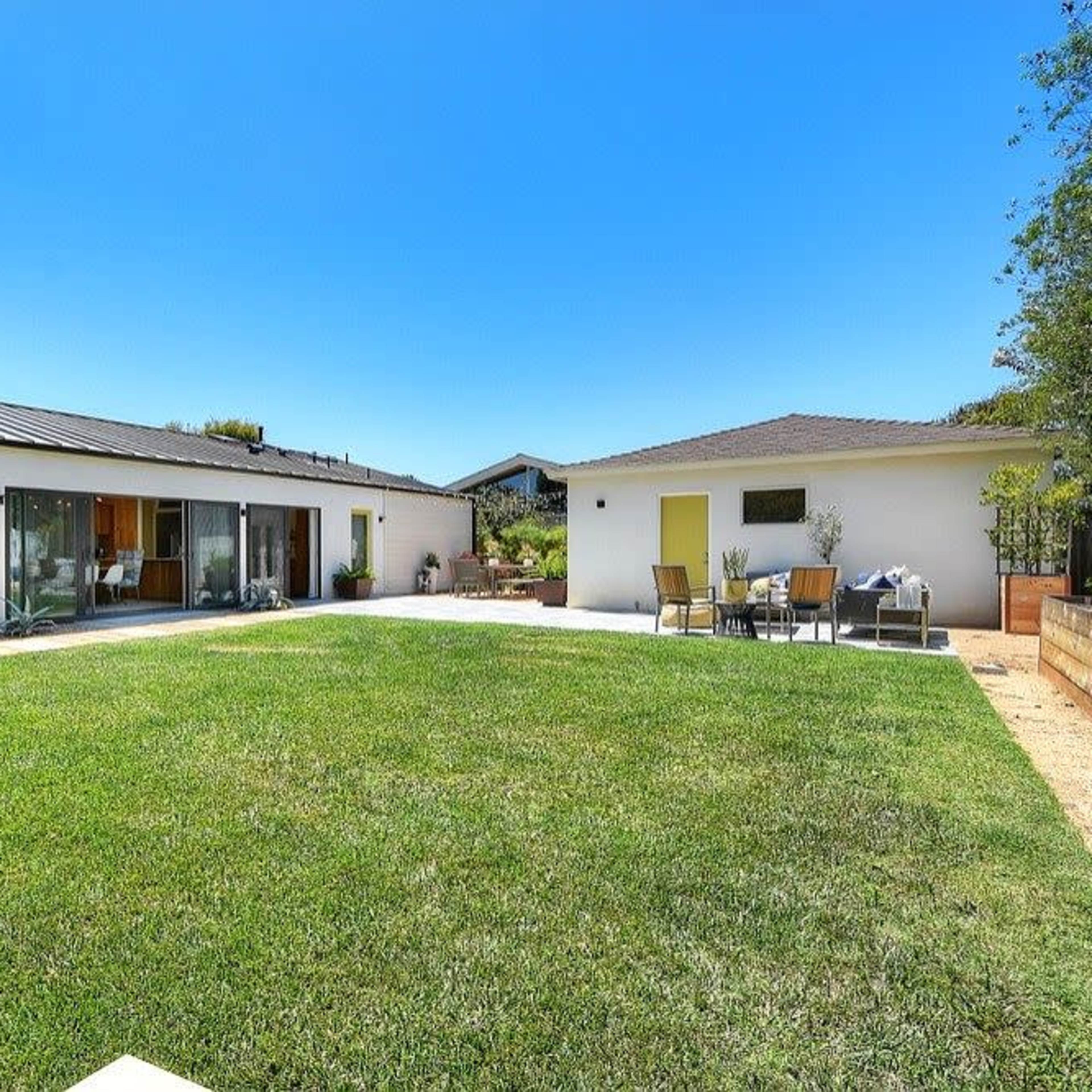 A modern single-story house with large glass sliding doors opens to a spacious backyard featuring a well-maintained lawn and patio furniture.