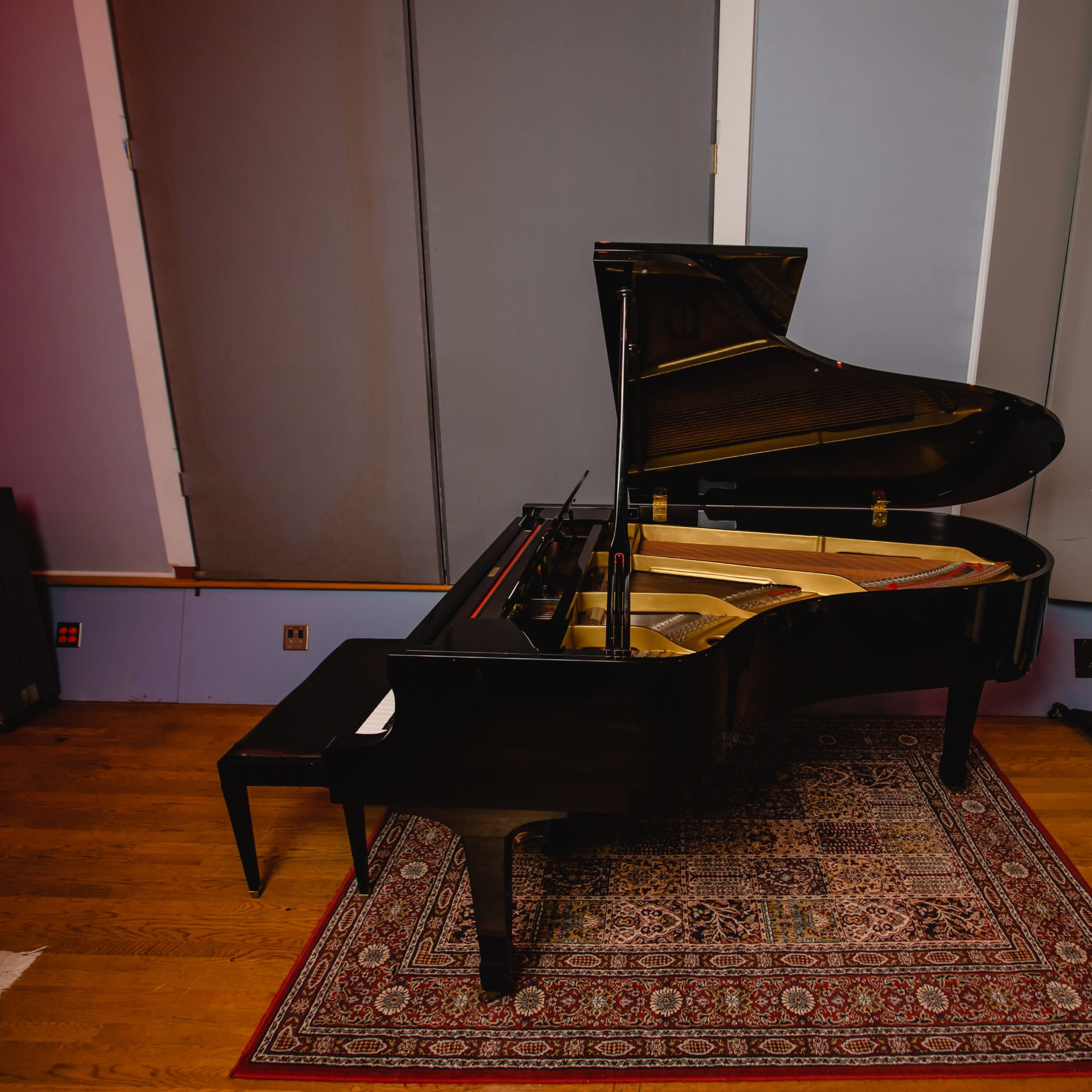 A black grand piano is positioned on a wooden floor, accented by a patterned rug, in a sound studio with muted wall colors.