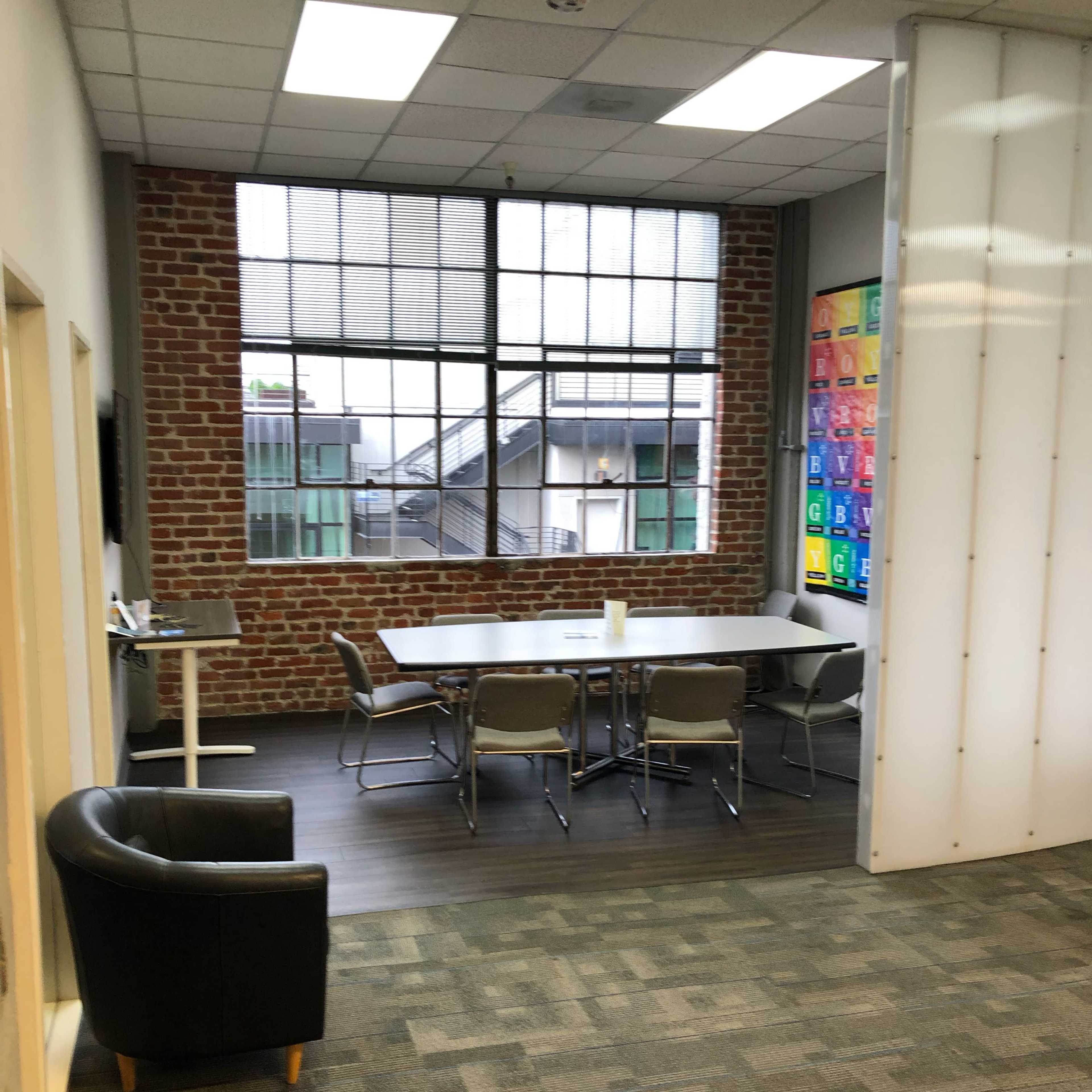A meeting room with a rectangular table surrounded by chairs, a brick wall, large windows, and a separate seating area featuring a black chair.