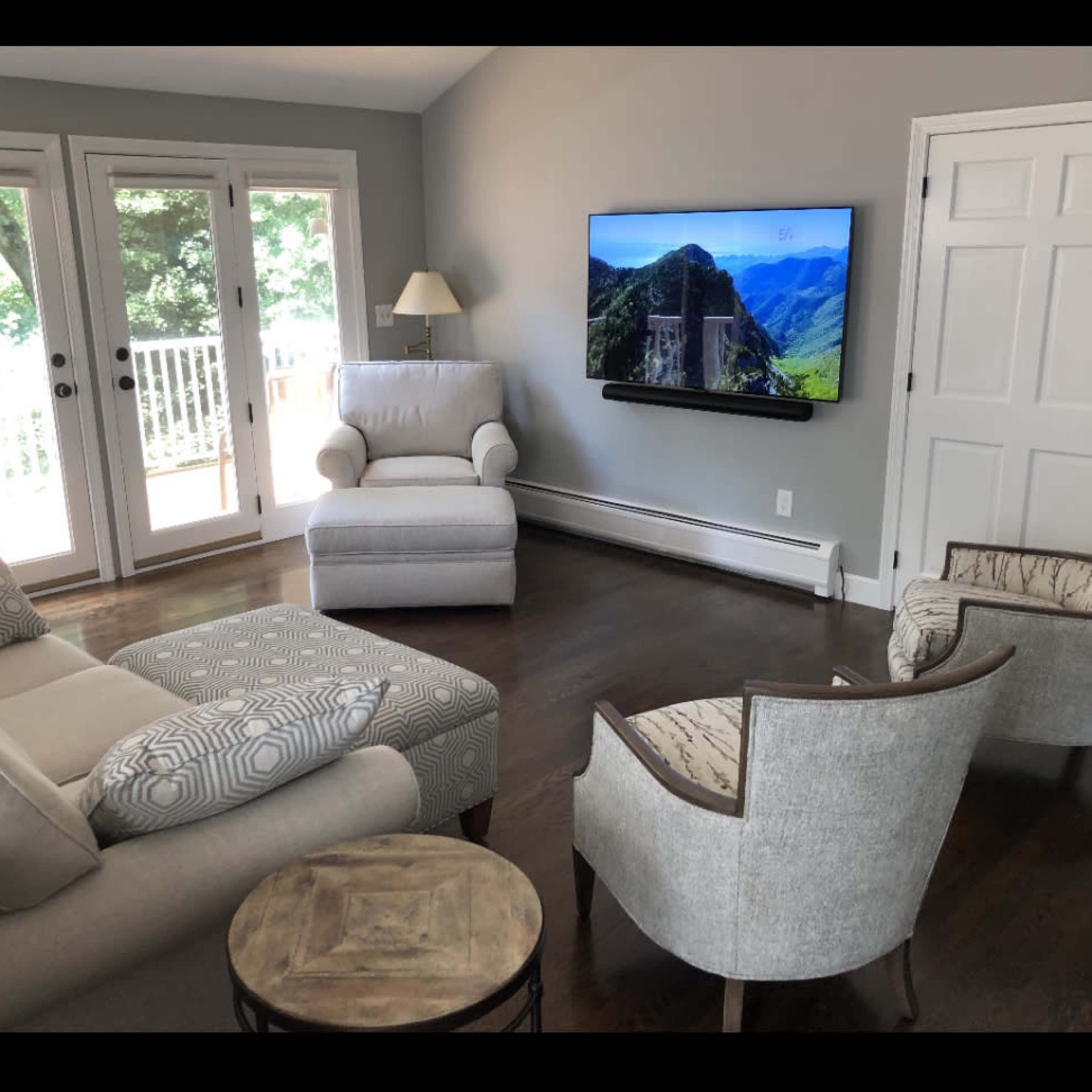 A living room furnished with a light-colored armchair, two upholstered chairs, a sectional sofa, a round table, and a wall-mounted television, with large windows allowing natural light.