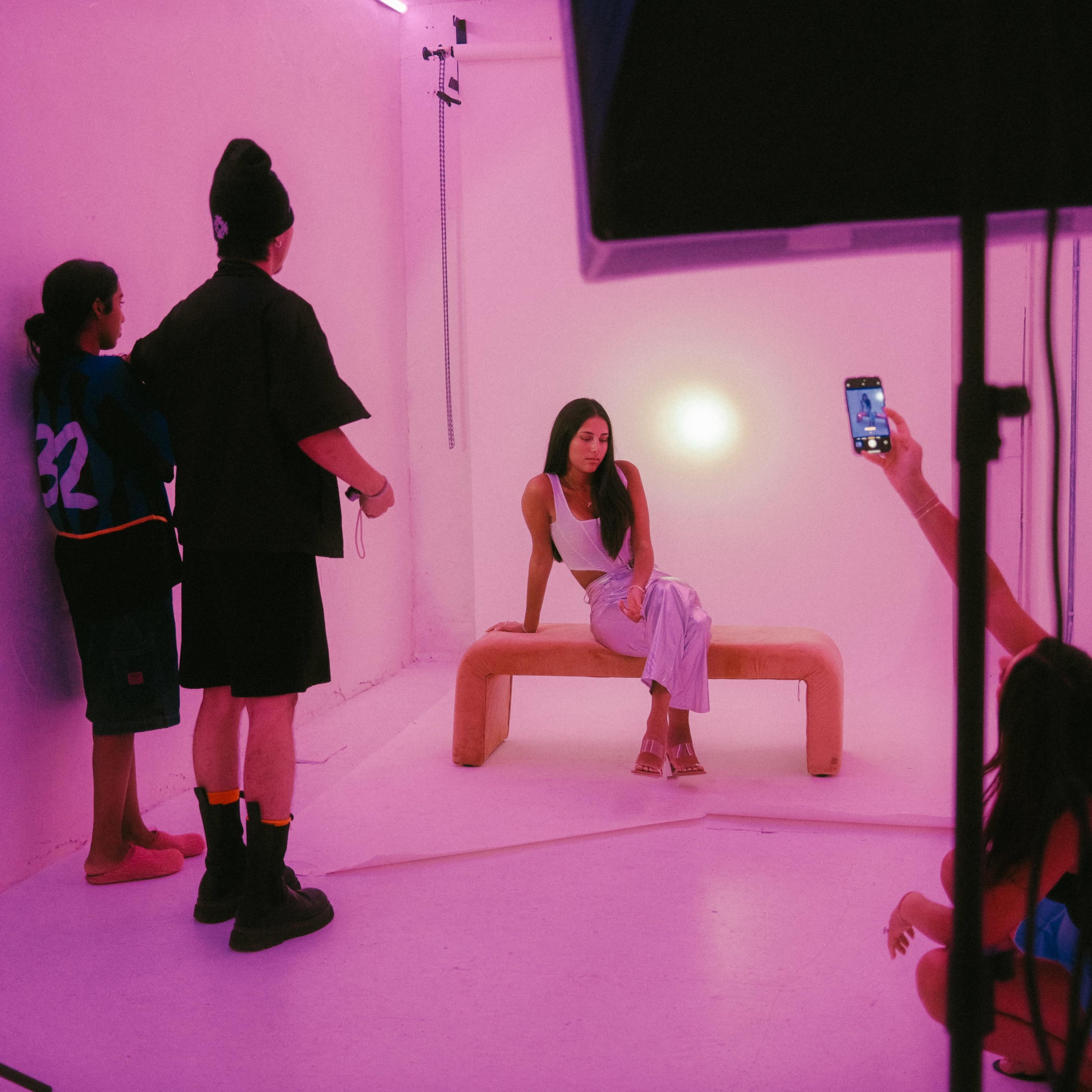 A model sits on a curved bench in a brightly lit studio while a group of people engage around her, with one person taking a photo.
