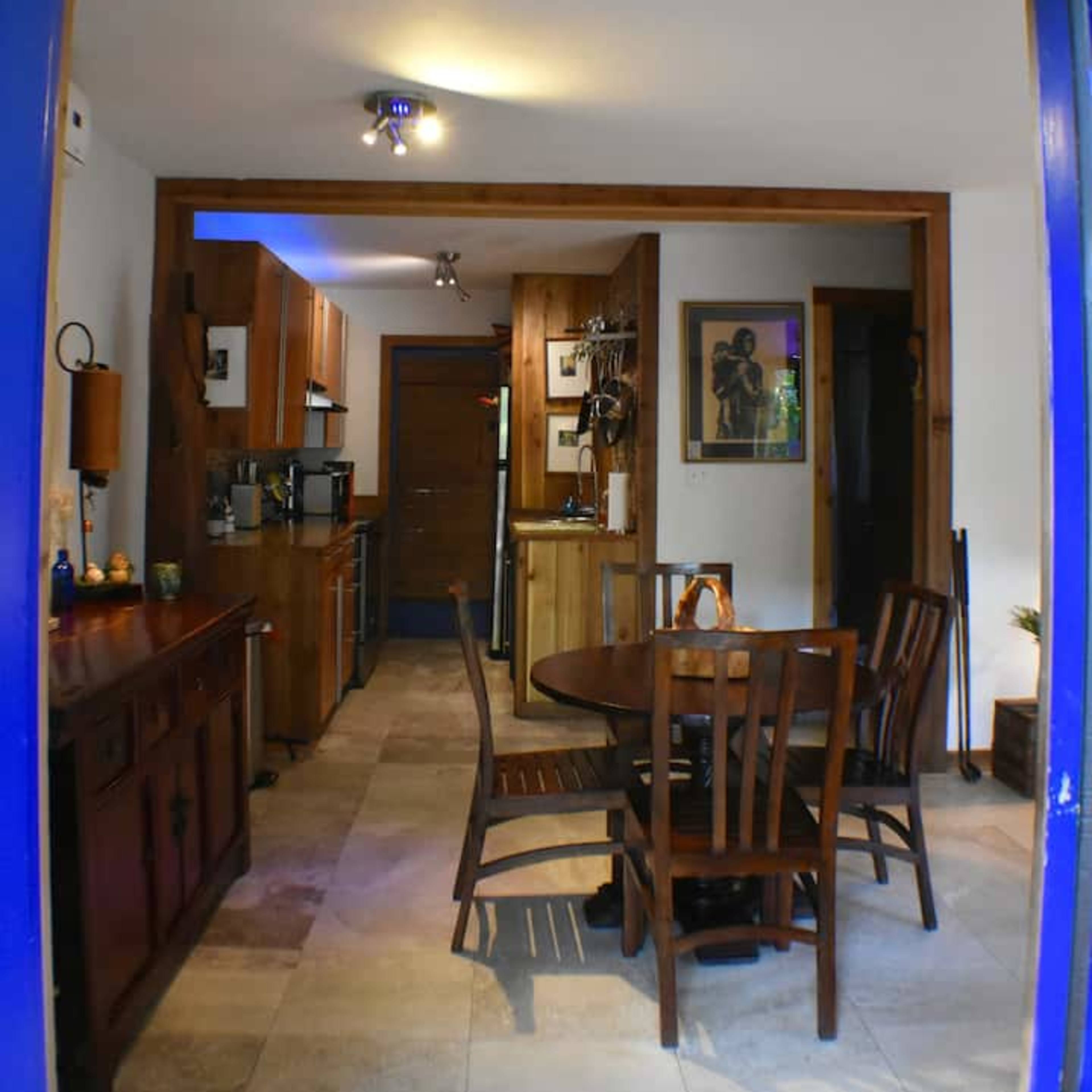 A kitchen with wooden cabinets, a circular dining table, and tiled flooring, viewed through a blue-framed doorway.