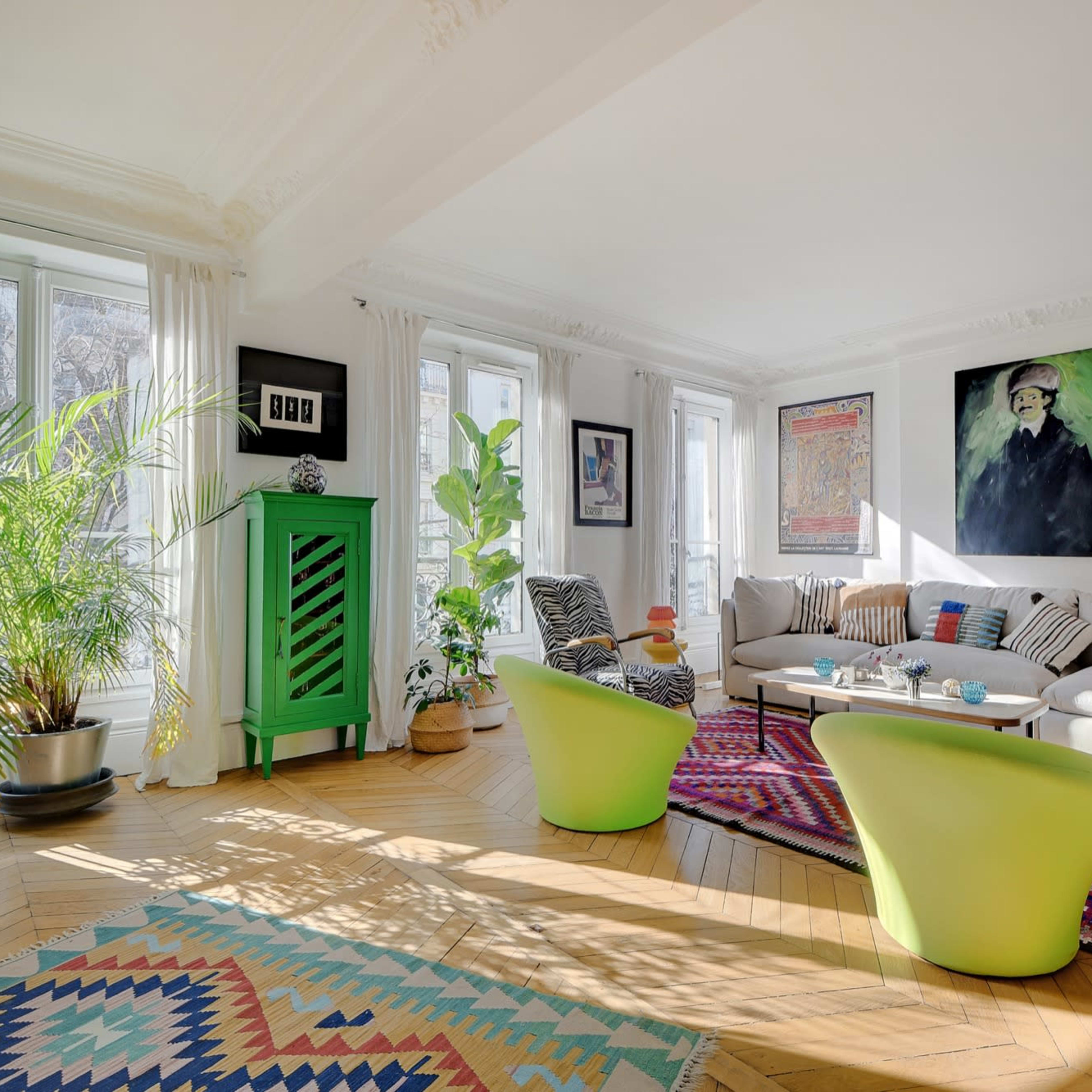 A brightly lit living room featuring a green cabinet, a colorful rug, a sofa with various cushions, and large windows with plants.
