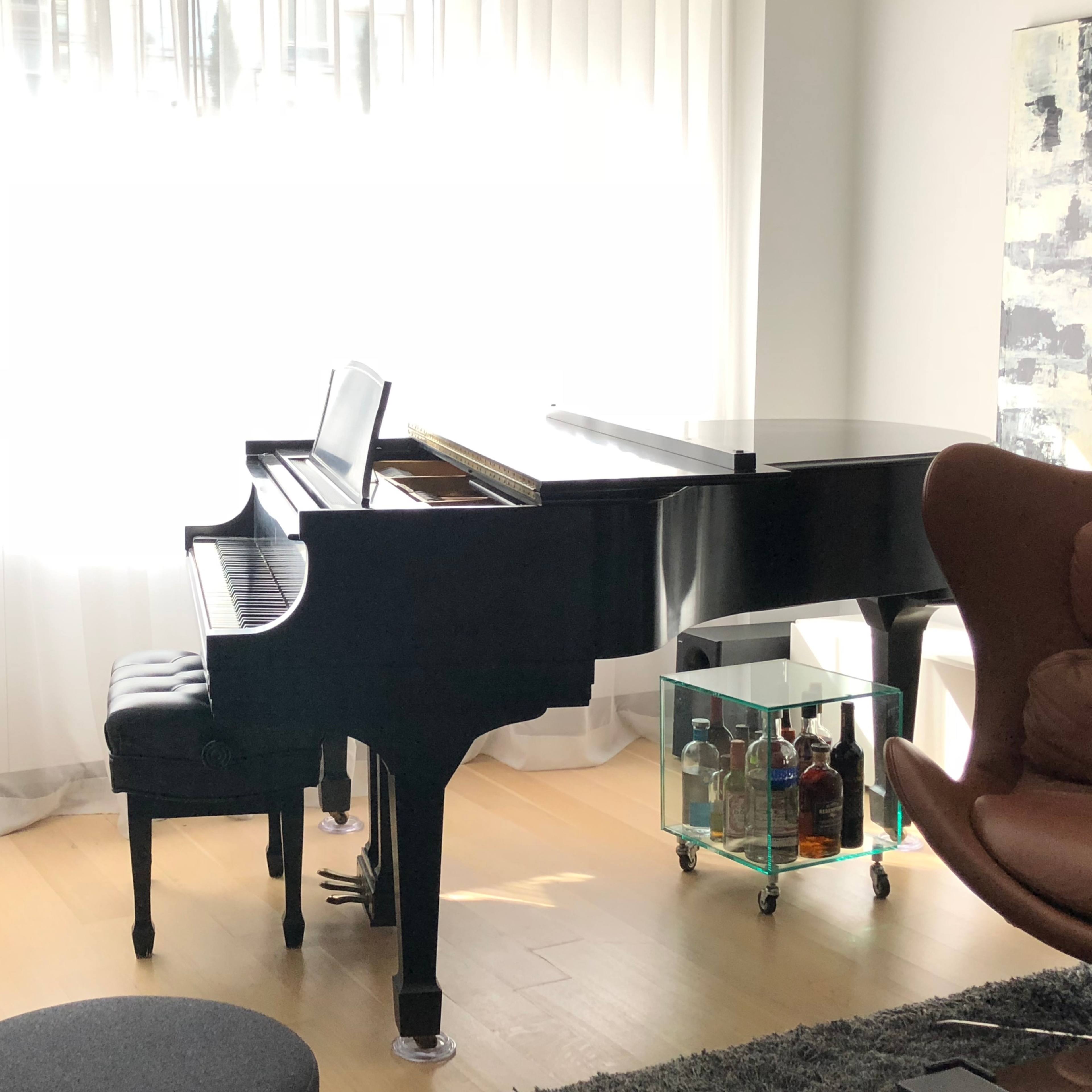 A black grand piano sits beside a window, with a stool in front and a glass cart holding bottles nearby.