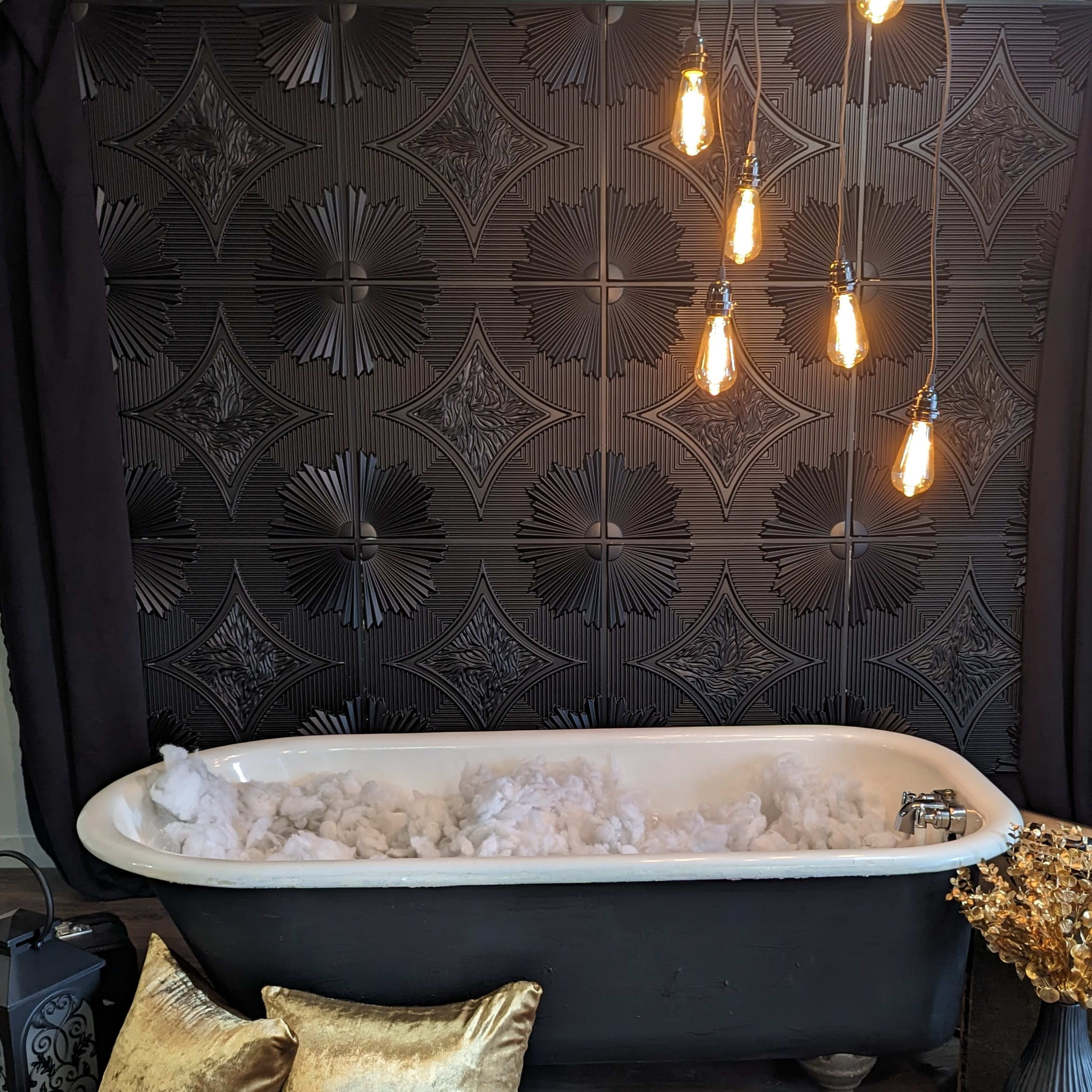 A vintage bathtub filled with white fluff sits against a patterned black wall, illuminated by hanging light bulbs, surrounded by decorative pillows and a plant.