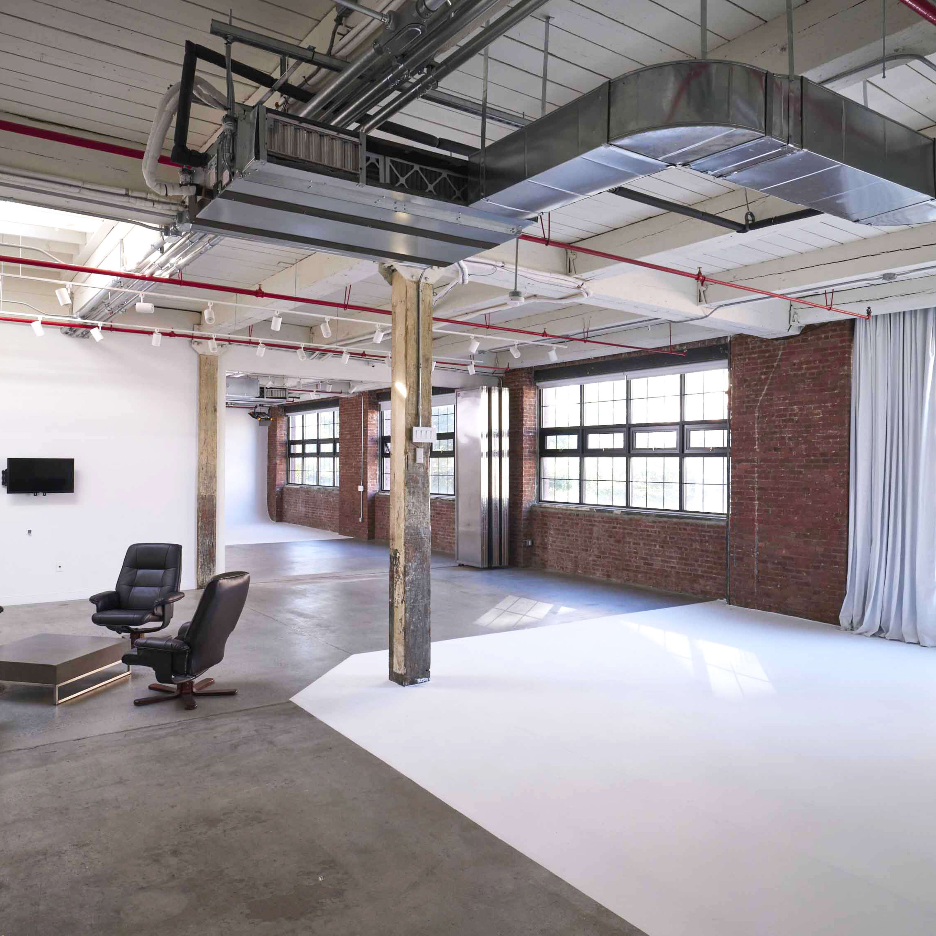 An industrial-style interior space with concrete floors, exposed beams, and large windows, featuring a small seating area with two black armchairs and a wooden coffee table.