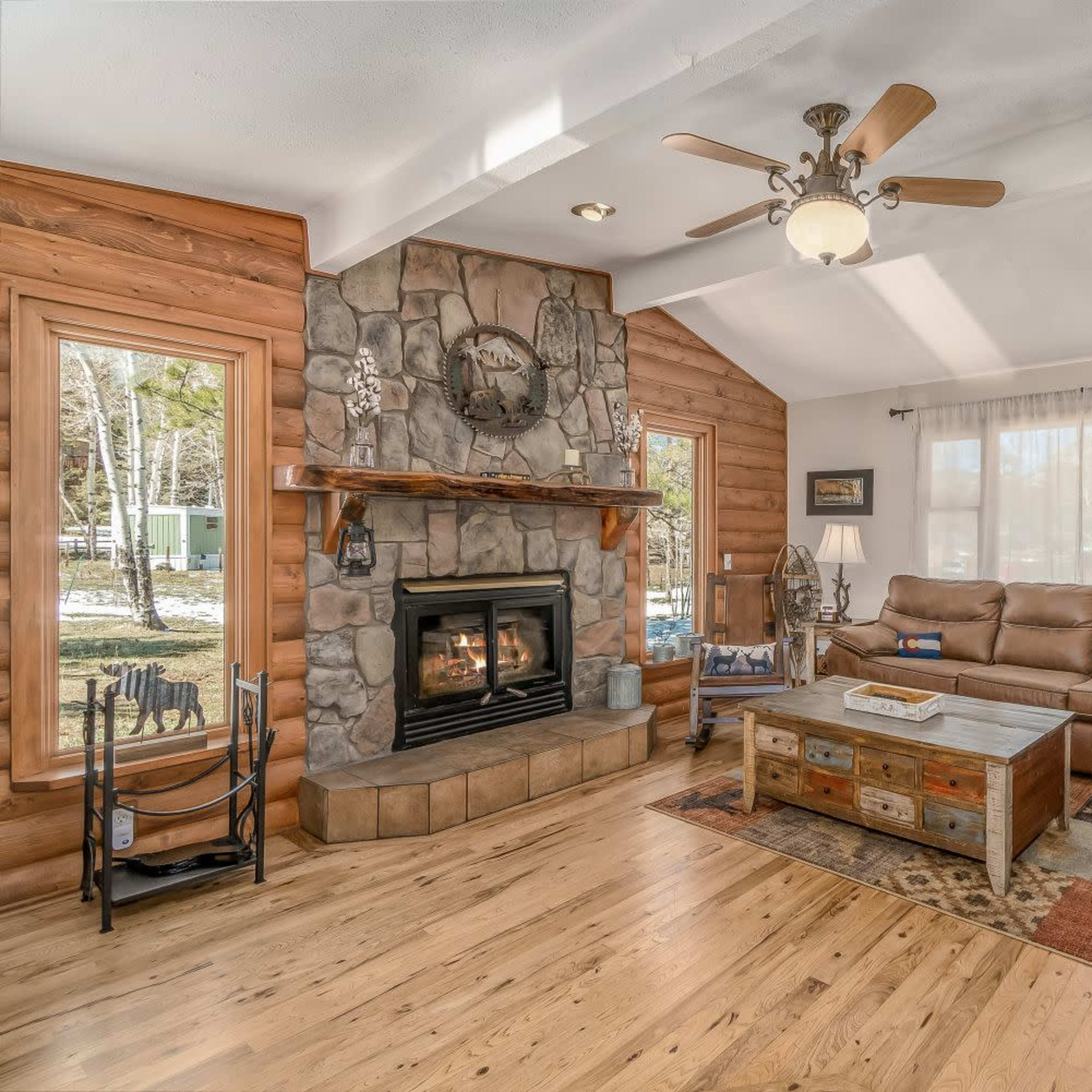 A cozy living room with a stone fireplace, large windows, a sectional sofa, and wooden flooring.