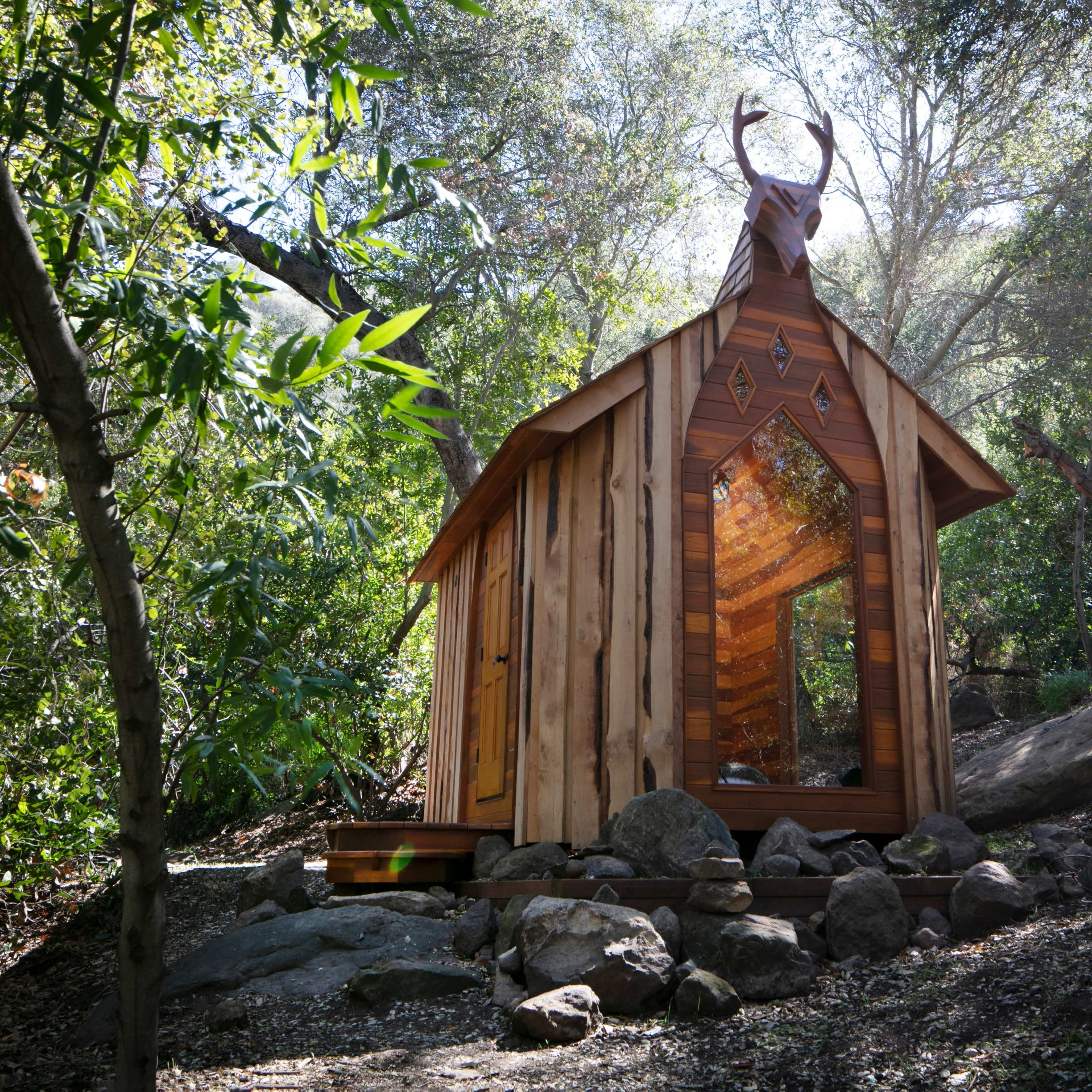 A small wooden cabin with a peaked roof and deer antlers on top is nestled among trees and rocks in a forested area.