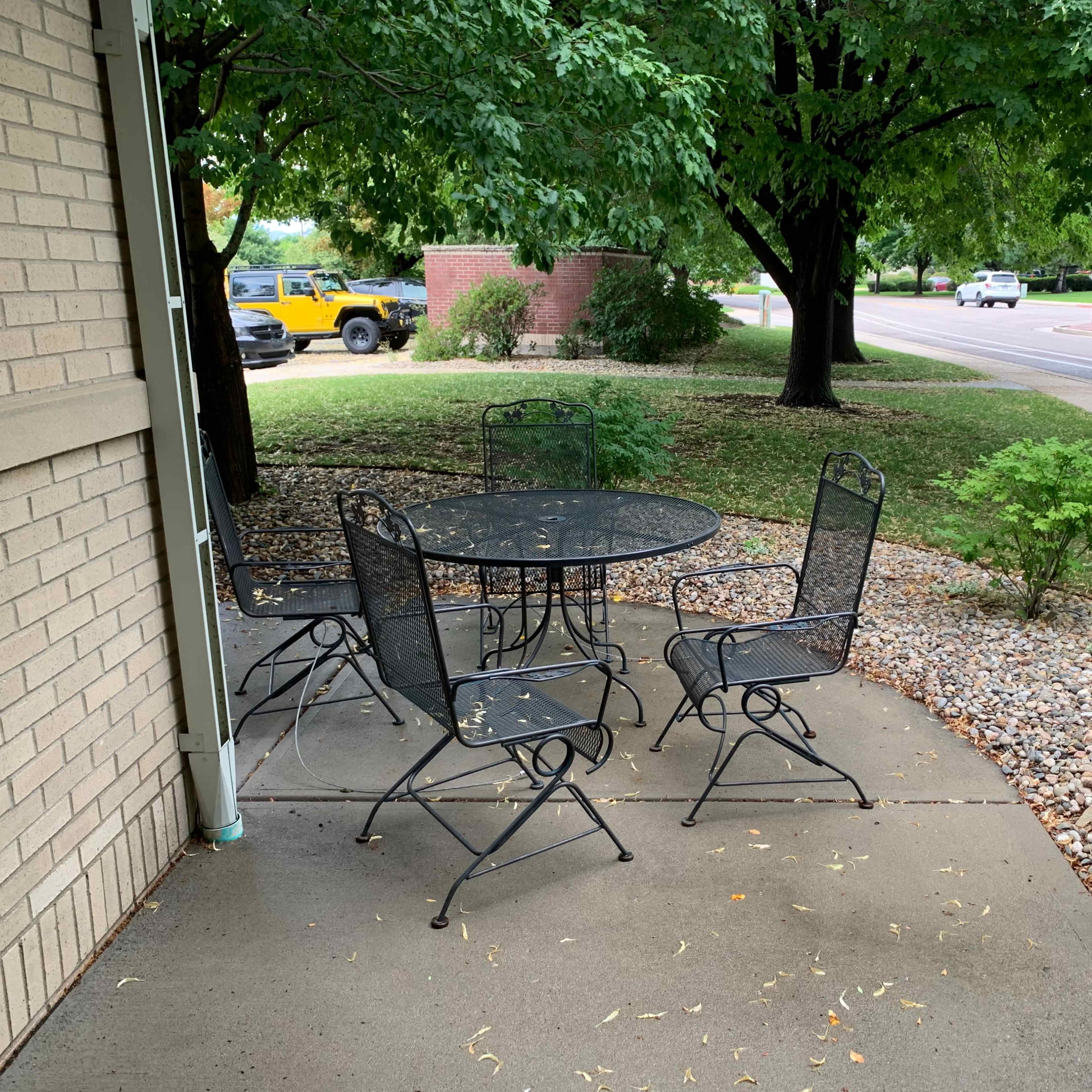 A black metal table with four chairs is positioned on a concrete patio surrounded by gravel and greenery near a street.