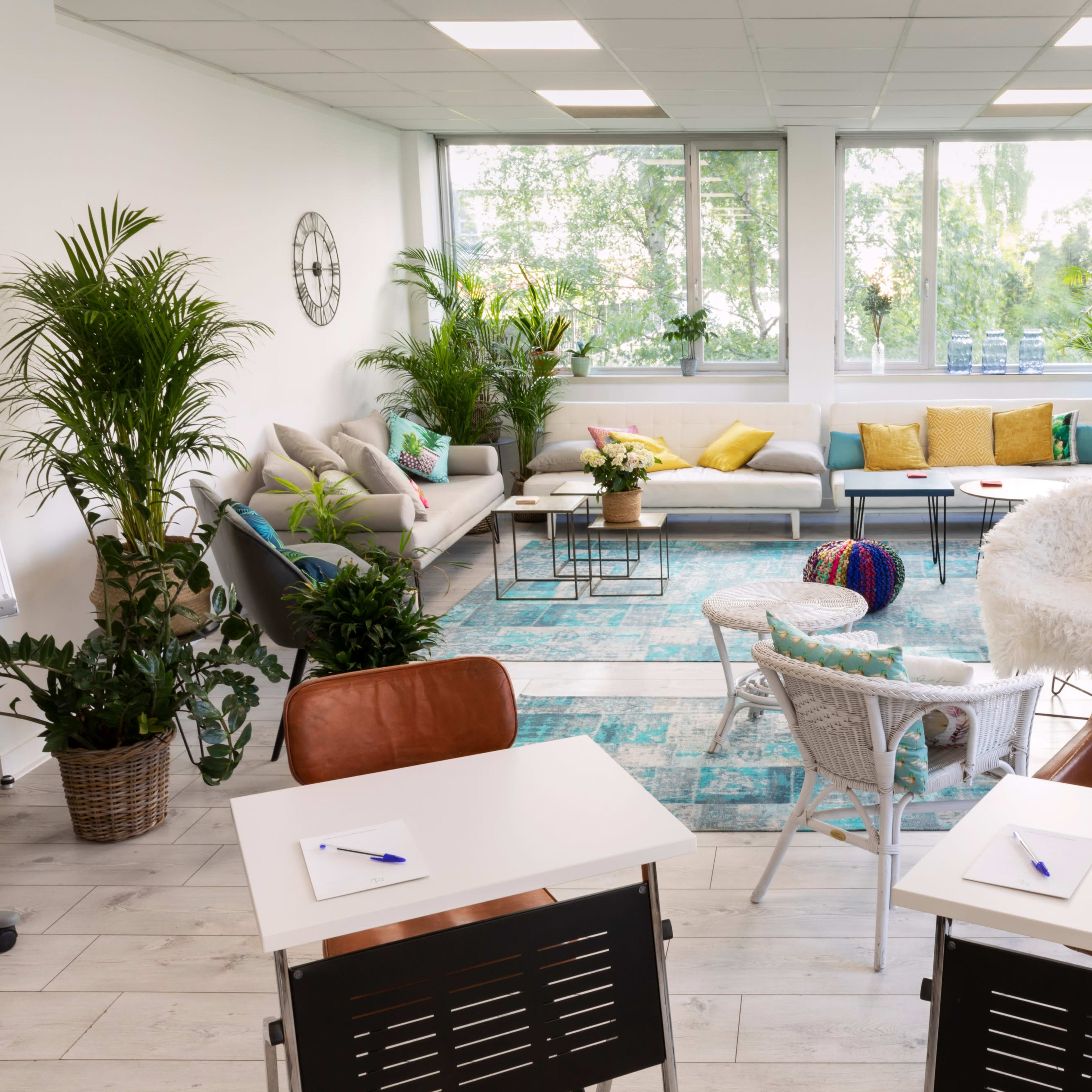 A bright, modern workspace featuring comfortable seating areas, green plants, and two desks with pens on them.