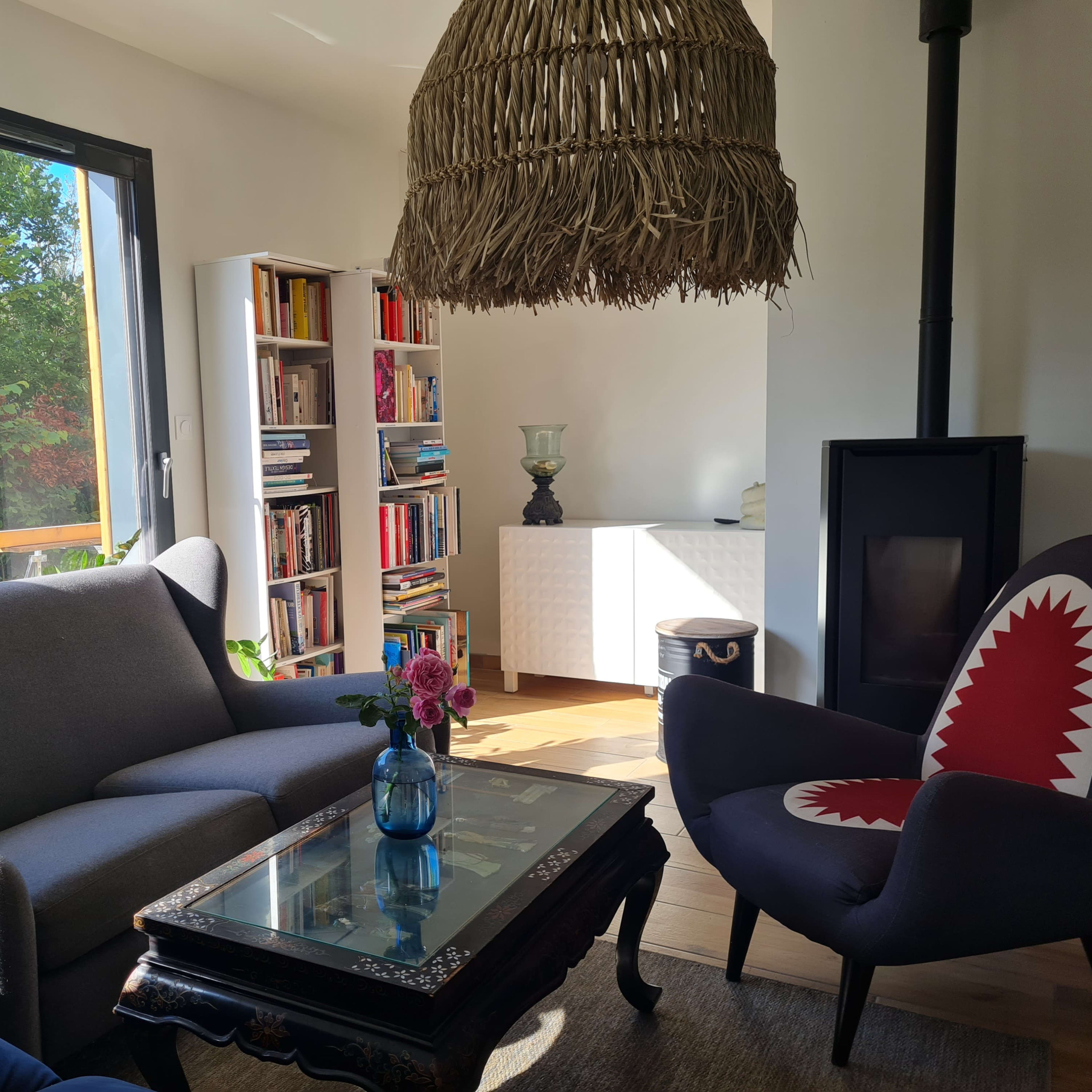 A cozy living room with a large window, showcasing a bright outdoor space, a bookshelf filled with books, and two distinct chairs arranged around a coffee table.