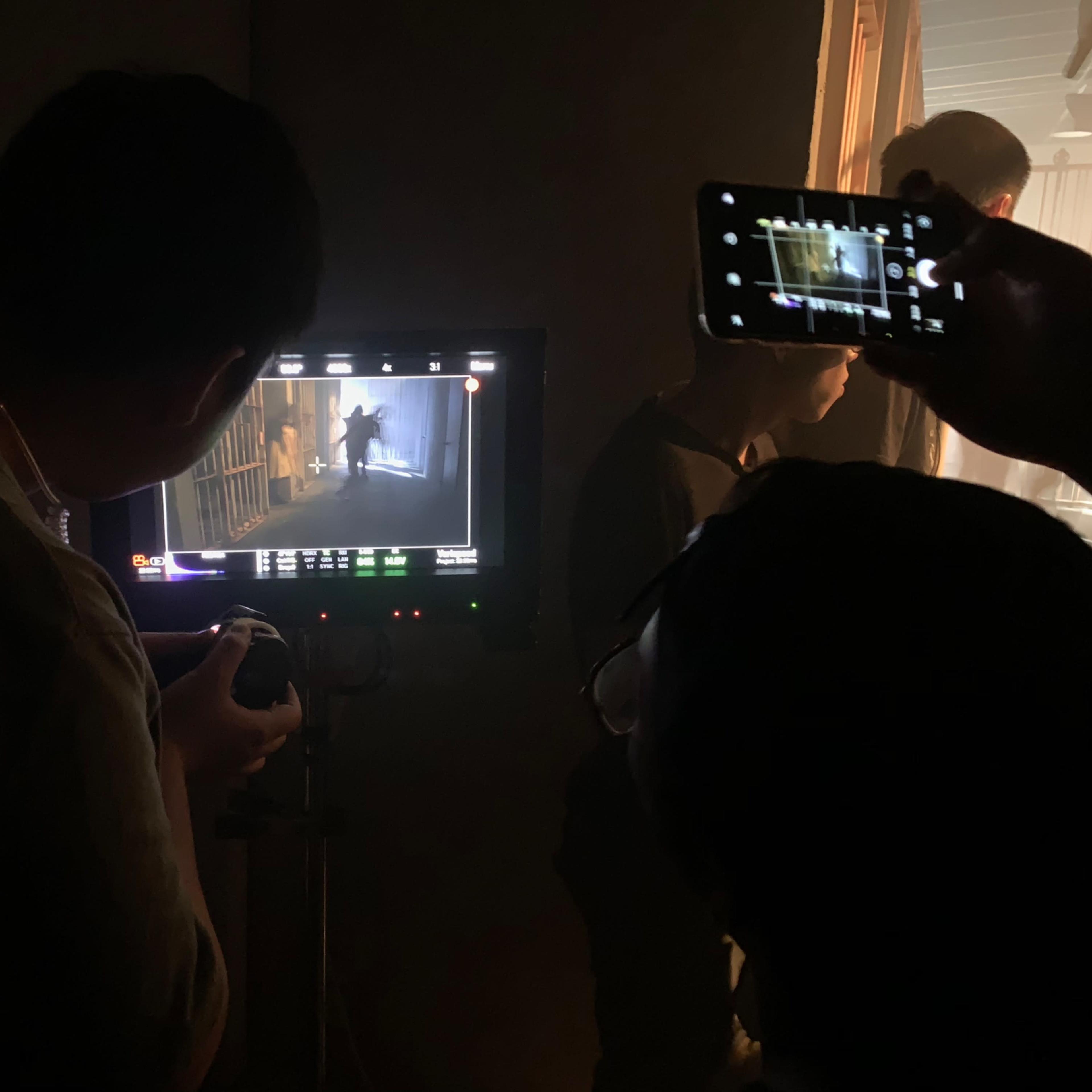A group of people is observing a scene being filmed, with one person holding a camera and another using a phone to record the monitor displaying the footage.